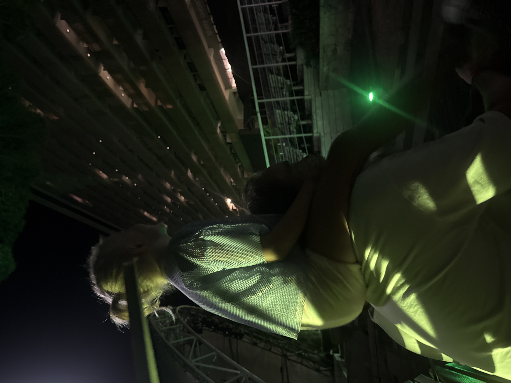 In the city of Yalta, Ukraine, a person is seen from behind, carrying a young child on their shoulders. The child, with blonde hair in a ponytail, wears a light-colored mesh shirt and shorts. They are looking off to the right. The person carrying them is wearing a light-colored t-shirt, illuminated by green lighting that casts strong shadows. In the background, a large, multi-story building with many illuminated windows is visible, suggesting an urban or resort setting at night. There are also metal structures and seating arrangements, possibly part of an outdoor venue or amphitheater. A bright green light beam is visible in the lower right portion of the image, adding to the atmospheric lighting.