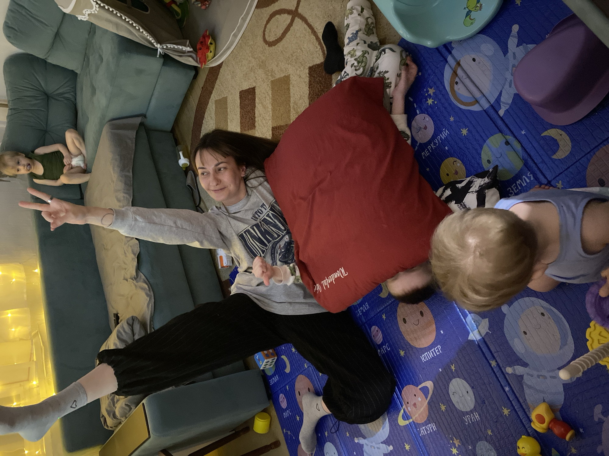 Scene depicting a woman lying on a blue planet-themed play mat with two children, likely in a domestic setting, though the location in Votkinsk, Russia, is not verifiable from the image. The woman is lying on her back, making a peace sign with her left hand. She wears a gray sweatshirt and black pants. A large red pillow is positioned near her head and shoulder. One child, partially obscured by the pillow, appears to be lying prone. Another child, wearing a blue shirt, is positioned to the right of the woman, looking down at something on the play mat. The play mat displays images of planets and astronauts. To the left, there is a green sectional couch with a child lying on it. The background includes a patterned rug and a play tent. The overall lighting is warm and diffused.