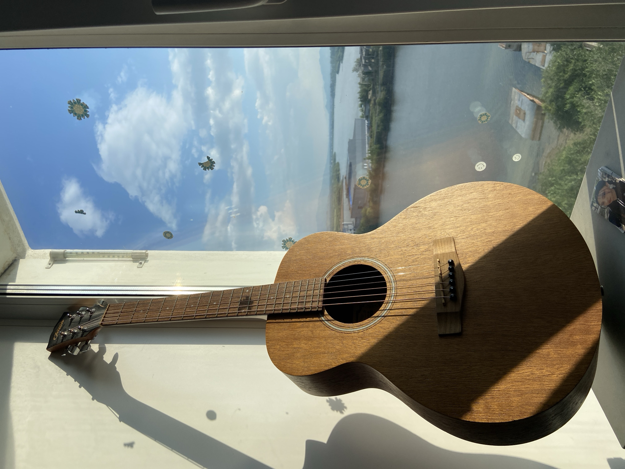 The image shows an acoustic guitar leaning against a white wall next to a window. The window offers a view of a river, with some buildings and mountains in the distance. The sun is casting a shadow of the guitar on the wall. There are a few decorative stickers on the window. The overall scene appears to be indoors, likely an apartment or studio, in Ulan-Ude, Russia, given the landscape and architecture visible through the window.