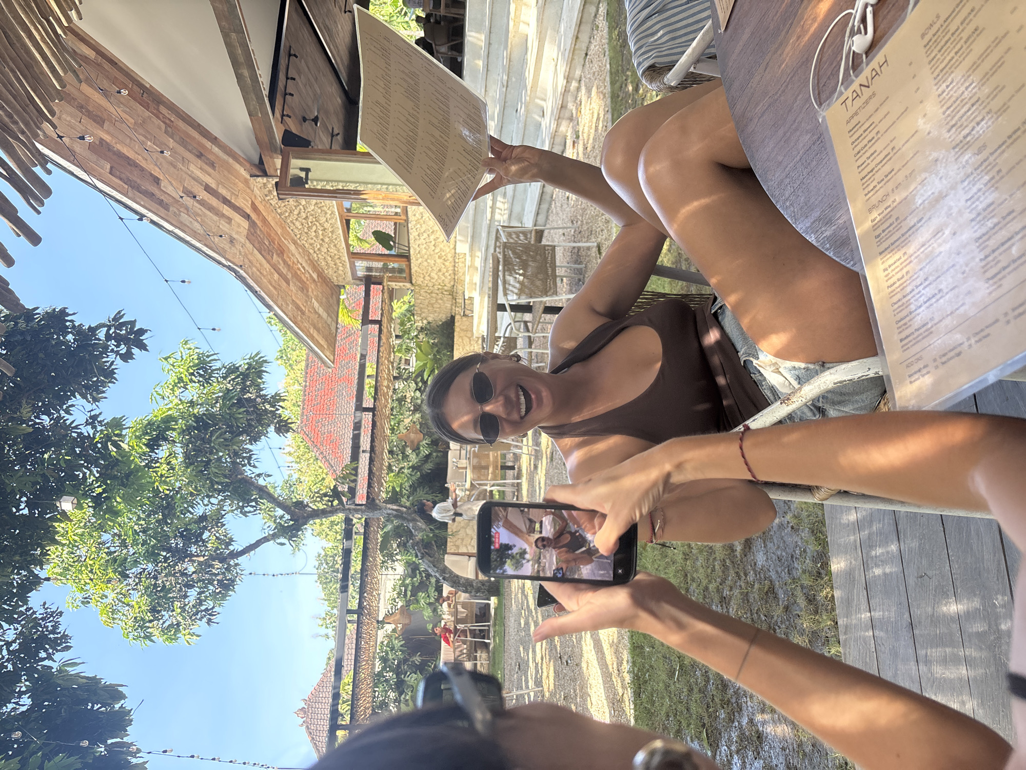 Captured outdoors in Kuta Selatan, Indonesia, the image shows a sunlit scene with lush foliage and architectural elements visible in the background, including tiled roofs. The image centers on a woman with sunglasses holding a menu card, facing the camera with a smile. Another individual is taking a picture of her with a smartphone. A menu with the word "TANAH" prominently displayed lies on the table. The environment is relaxed, with a clear sky peeking through the trees, indicating a bright, daytime setting.