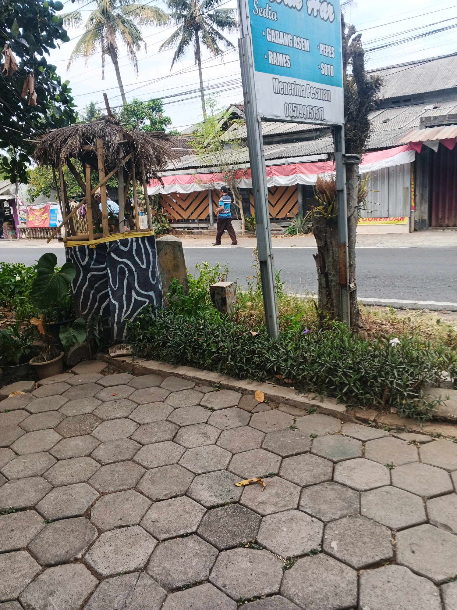 The image captures a street scene in Ungaran, Indonesia, likely a commercial area given the signage and buildings. In the foreground, hexagonal paving stones form a walkway. To the left, a small, rustic structure with a thatched roof stands amidst lush green foliage. This structure appears to be decorated with a black and white abstract pattern on its lower section. Behind this, a blue and white sign is prominently displayed, listing various food items available, such as "GARANG ASEM", "PEPES", "RAMES", and "SOTO", along with a phone number for orders. A person in a blue and white striped shirt and brown pants is walking away from the camera towards the right side of the image, in front of a row of shops. These shops have red and white striped awnings and their facades suggest a simple, functional design. Palm trees are visible in the background, indicative of the tropical climate. The overall impression is that of a typical Indonesian street scene, with elements of local commerce and everyday life.
