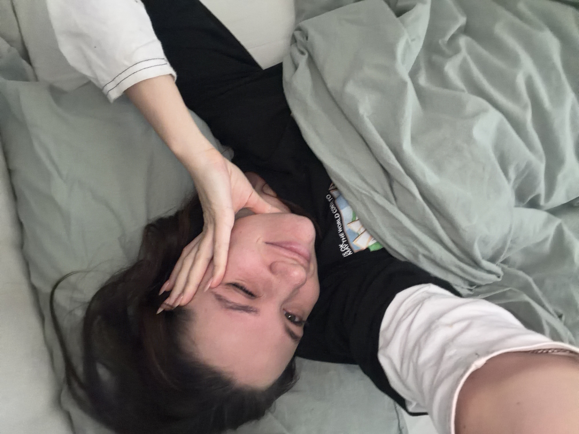 A young woman with long brown hair lies in bed, partially covered by a gray blanket. She wears a black long-sleeved shirt over a white undershirt. Her left hand is raised to her forehead, covering part of her face. The scene is lit by natural light. There is no information in the image to indicate the location is in Belgrade, Serbia.