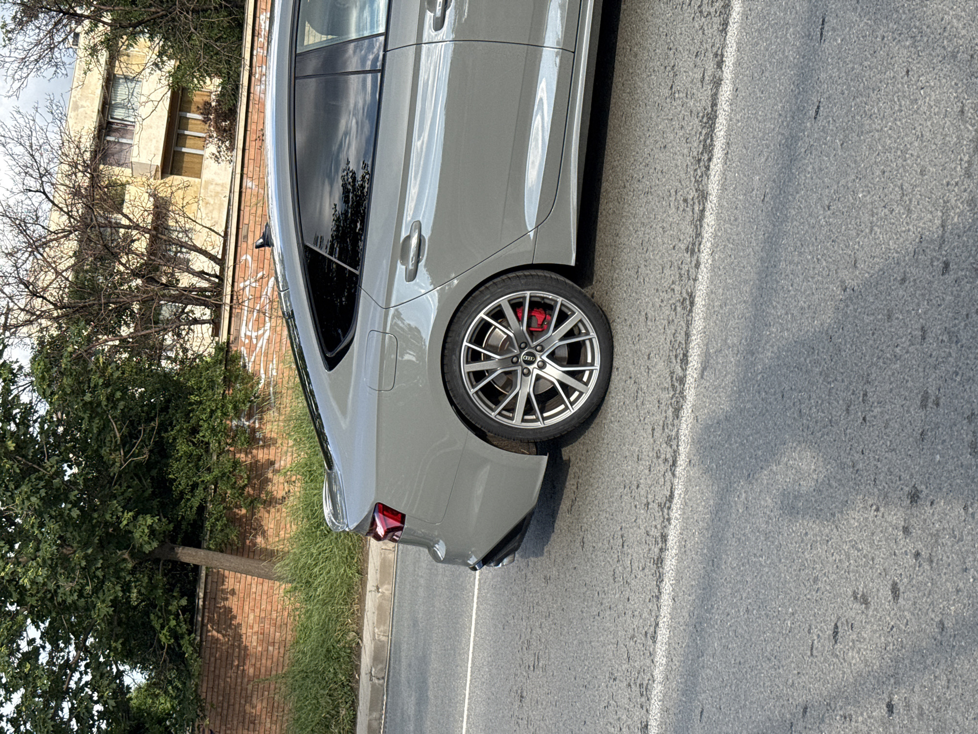 The image, taken in Sofia, Bulgaria, presents a horizontal view of the rear and side profile of a grey Audi car, likely a sportback or station wagon, parked or stopped by the side of an asphalt road during the daytime.
The car is the central subject, displaying its sleek, reflective grey paintwork and tinted windows. Prominent multi-spoke alloy wheels reveal bright red brake calipers behind them, indicating a performance-oriented vehicle. The Audi rings logo is visible on the center cap of the rear wheel. Reflections on the car's bodywork and windows suggest a bright, possibly partly cloudy sky.
To the left of the car, a red brick wall, lightly defaced with indistinct graffiti, runs along the roadside. Beyond the wall, an older, light-colored building with several rectangular windows is partially visible, suggesting an urban residential or commercial structure. Various trees, some with bare branches and others with green leaves, are interspersed between the wall and the building, partially obscuring them.
On the right side of the car, an asphalt road with a clear white lane marking extends into the distance. A strip of green grass borders the road on the far right, alongside a concrete curb.
The scene is well-lit by natural sunlight, casting shadows from the car and trees, consistent with a clear day. No people or specific activities are visible; the scene depicts a static moment in an urban or suburban environment.