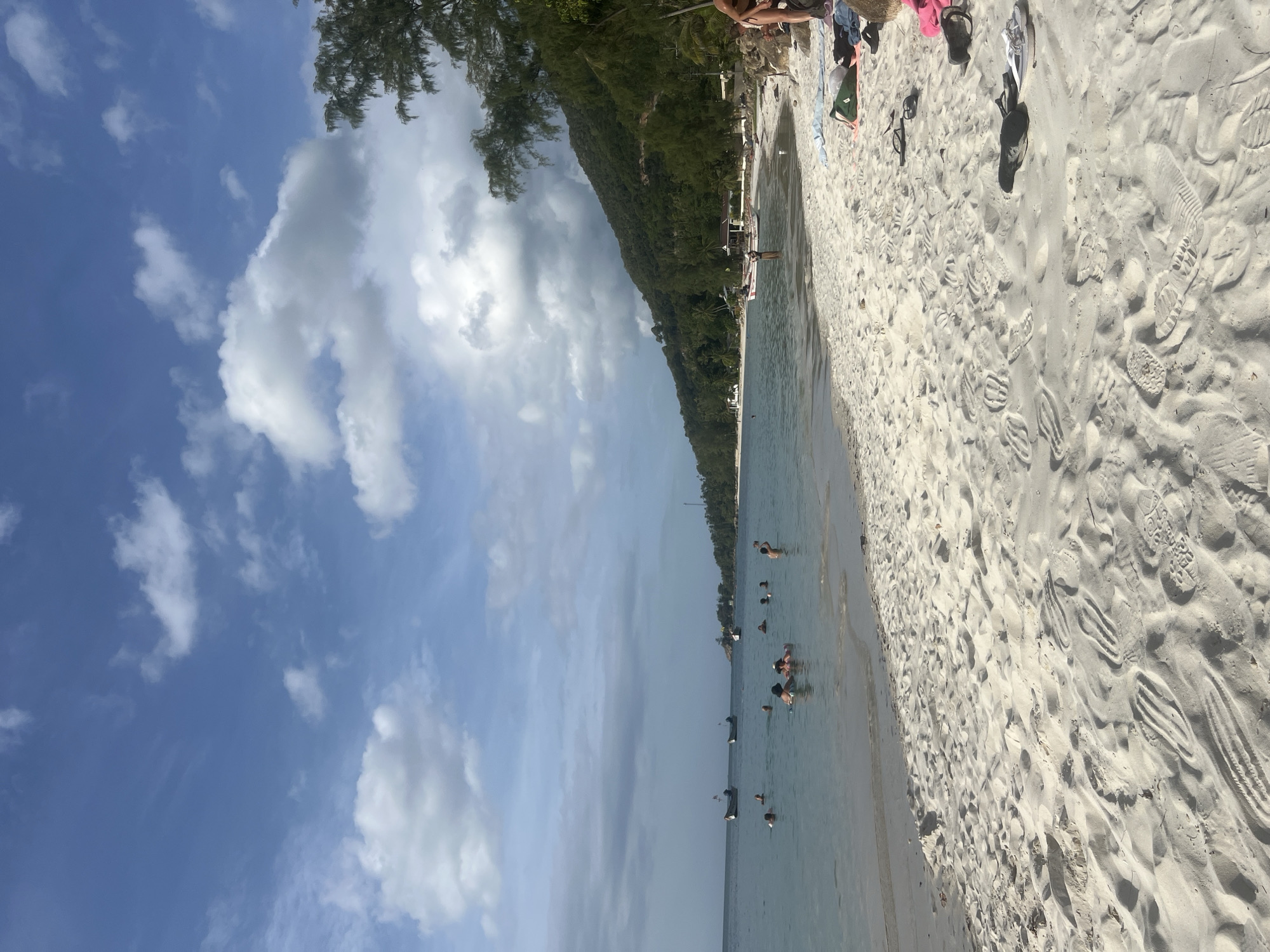 The image depicts a sandy beach with calm, turquoise waters under a partially cloudy blue sky. Several people are visible swimming and wading in the shallow sea, while some boats are anchored offshore. The beach is covered in footprints, suggesting recent activity. To the right, a lush green hillside rises steeply, dotted with trees and some buildings. In the foreground, beach items such as towels and flip-flops are scattered on the sand. The scene conveys a relaxed, summery atmosphere.