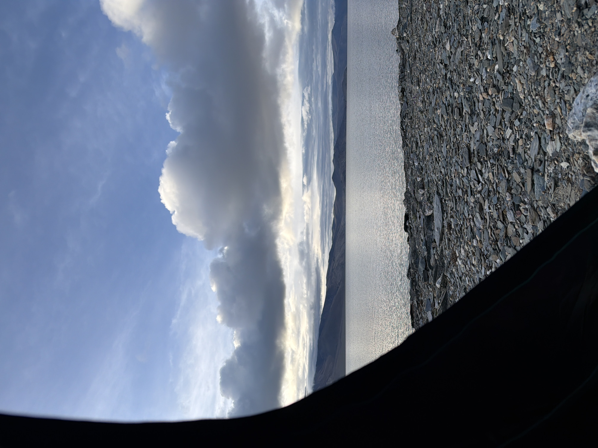 A wide-angle shot captures a serene natural landscape framed by a dark, curved object on the left, possibly the edge of a tent. The foreground is a rocky shoreline, with numerous grey and brown stones scattered across the ground. Beyond the shore, a calm body of water reflects the sky, its surface rippled by a gentle breeze. In the distance, a range of low, dark mountains forms the horizon, their peaks softened by the atmospheric conditions. The sky above is a mix of light blue with scattered white and grey clouds, some appearing heavy and dark, suggesting an overcast or transitioning weather pattern. The overall impression is one of a tranquil, remote setting, likely in the Himalayan region of India given the terrain and the atmospheric haze often seen at higher altitudes. No people, animals, or man-made structures are immediately visible, emphasizing the natural beauty of the scene.