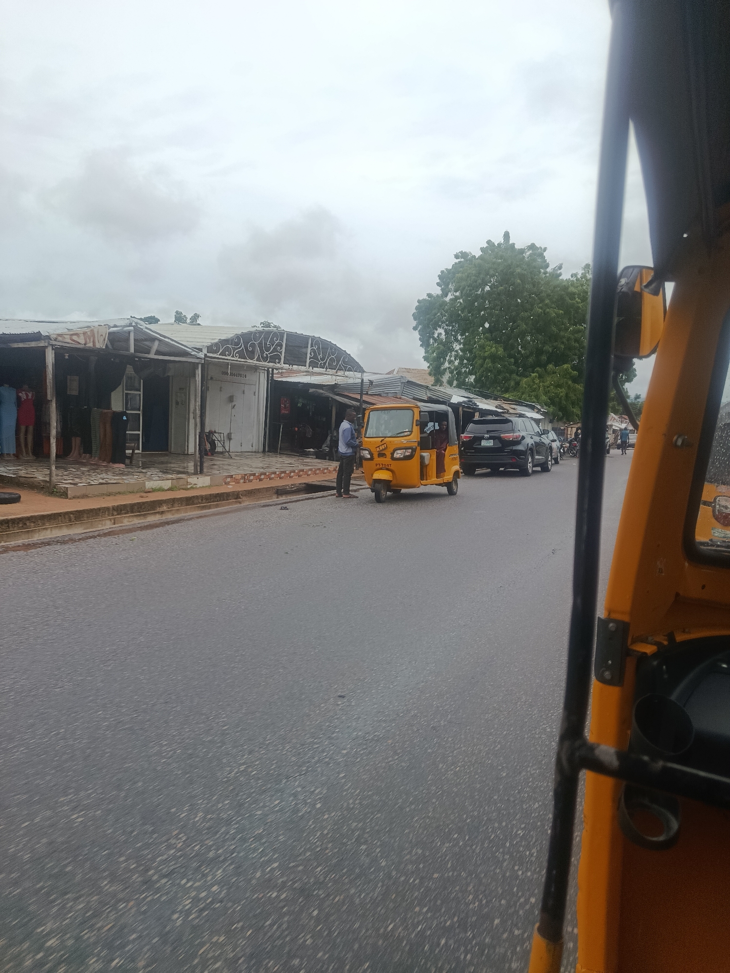 The image depicts a street scene in Jalingo, Nigeria, viewed from the perspective of a passenger inside a yellow auto-rickshaw. The auto-rickshaw is in the foreground on the right side of the frame, with its distinctive yellow body and black metal frame visible. In front of the auto-rickshaw, a black SUV is stopped on the right side of the road. Beyond the SUV, other vehicles, including motorcycles and what appears to be another auto-rickshaw, are visible further down the street.
On the left side of the road, a row of shops with corrugated metal roofs and open fronts line the street. Some shops display clothing on mannequins and hangers. Several people are visible in front of and within the shops. The road itself is paved and appears wet, suggesting recent rain. The sky is overcast with gray clouds. The environment is characteristic of a bustling urban or semi-urban area in Nigeria, with commercial activity and various modes of transportation. The shops have a simple, functional design, typical of roadside businesses in the region. The overall atmosphere is one of everyday life and commerce.