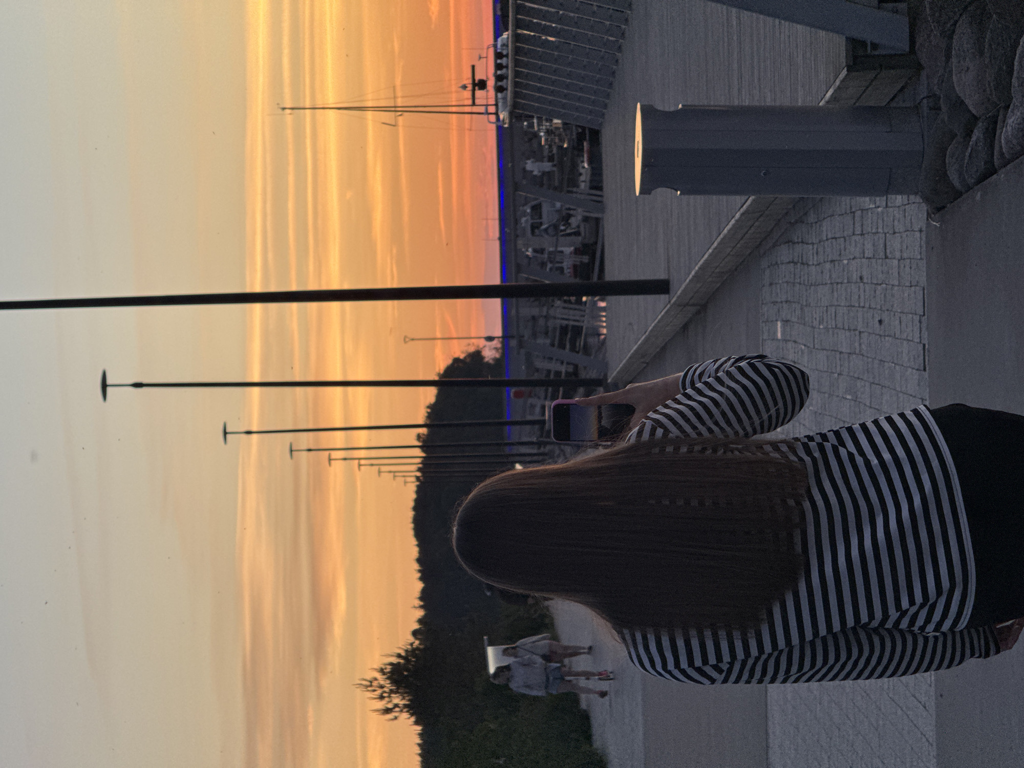 The image captures a sunset scene at a harbor in Tallinn, Estonia. In the foreground, a person with long, dark hair is seen from behind, wearing a black and white striped long-sleeved shirt and black pants. They are holding a smartphone in their right hand, appearing to take a photo or video of the sunset. The phone screen displays a vibrant orange and pink sky.
The background reveals a paved walkway and a wooden pier. Several lampposts are spaced along the pier. In the distance, a sailboat is docked in the harbor, and other boats are visible. The sky is filled with the warm hues of a sunset, with orange and yellow streaks dominating the horizon. On the left side of the image, two other individuals are walking on the path; one is on a scooter, and the other is walking behind. A line of trees forms a dark silhouette against the colorful sky. The overall atmosphere is serene and picturesque, typical of a coastal sunset in a city like Tallinn.