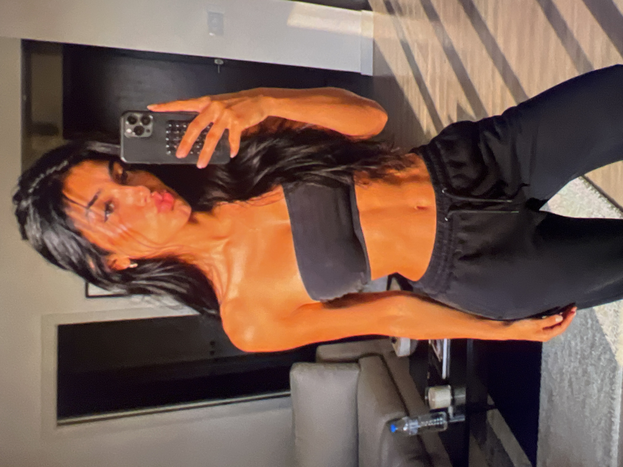 A young woman with dark hair and tan skin is taking a selfie in a room. She is wearing a black strapless top and black sweatpants. Her lips are pursed. In the background, there is a sofa, a coffee table with a water bottle and a candle, and a door. Sunlight is streaming in through blinds, casting striped shadows on the floor.