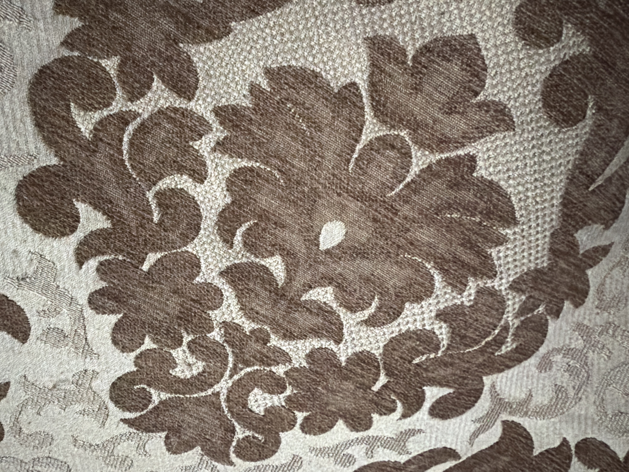 The image displays a close-up, overhead view of a textured fabric with a damask pattern. The fabric is predominantly beige with an intricate floral design in a darker brown color. The pattern features large, stylized flowers with layered petals and scrolling leaf-like elements. The texture of the fabric appears to be a woven material, possibly a tapestry or upholstery fabric, with a slightly raised nap in the brown areas creating a sense of depth. The lighting is even, highlighting the details of the pattern and the texture of the material. There are no people, animals, or specific environmental features visible in this close-up shot of the fabric. This image is not located in the city of Kaspiysk, Russia, as there are no discernible clues to suggest this location.