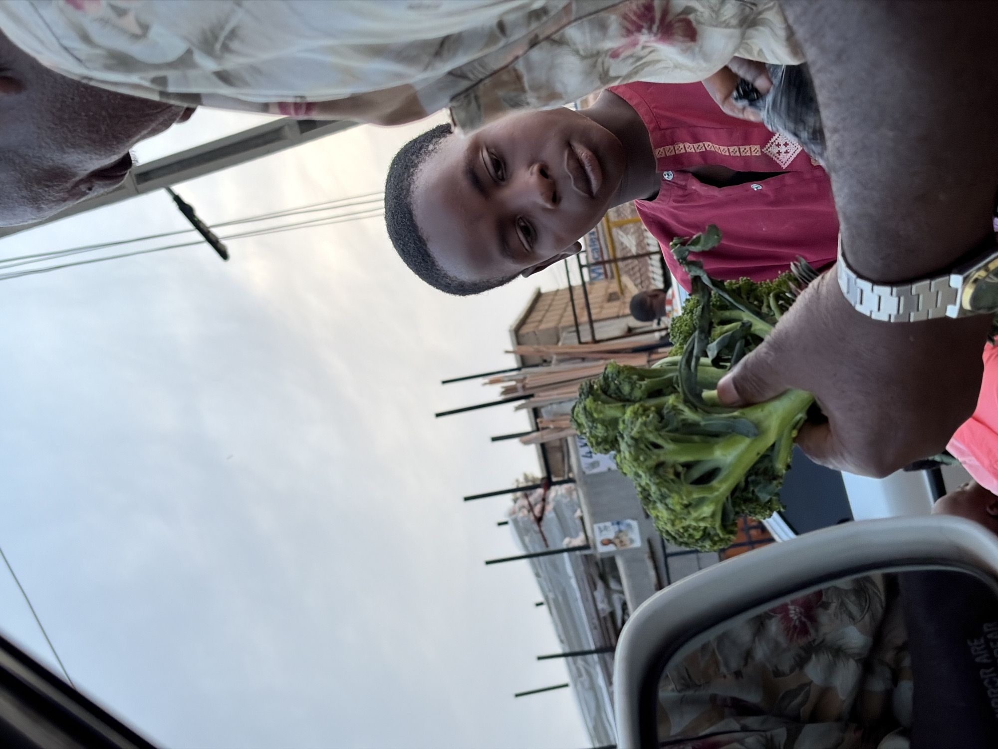 A low-angle shot taken from inside a vehicle shows a person holding out a head of broccoli for sale. The person offering the broccoli is standing beside the car, and the top of their head is visible. The head of broccoli is in focus. A young person in a red shirt looks at the camera. In the reflection of the car's side mirror, part of the interior of the car is visible, with floral patterned fabric. Through the car window, a building, possibly a shop, is visible, along with power lines. The sky is overcast. The location is Gura Top, Nigeria.