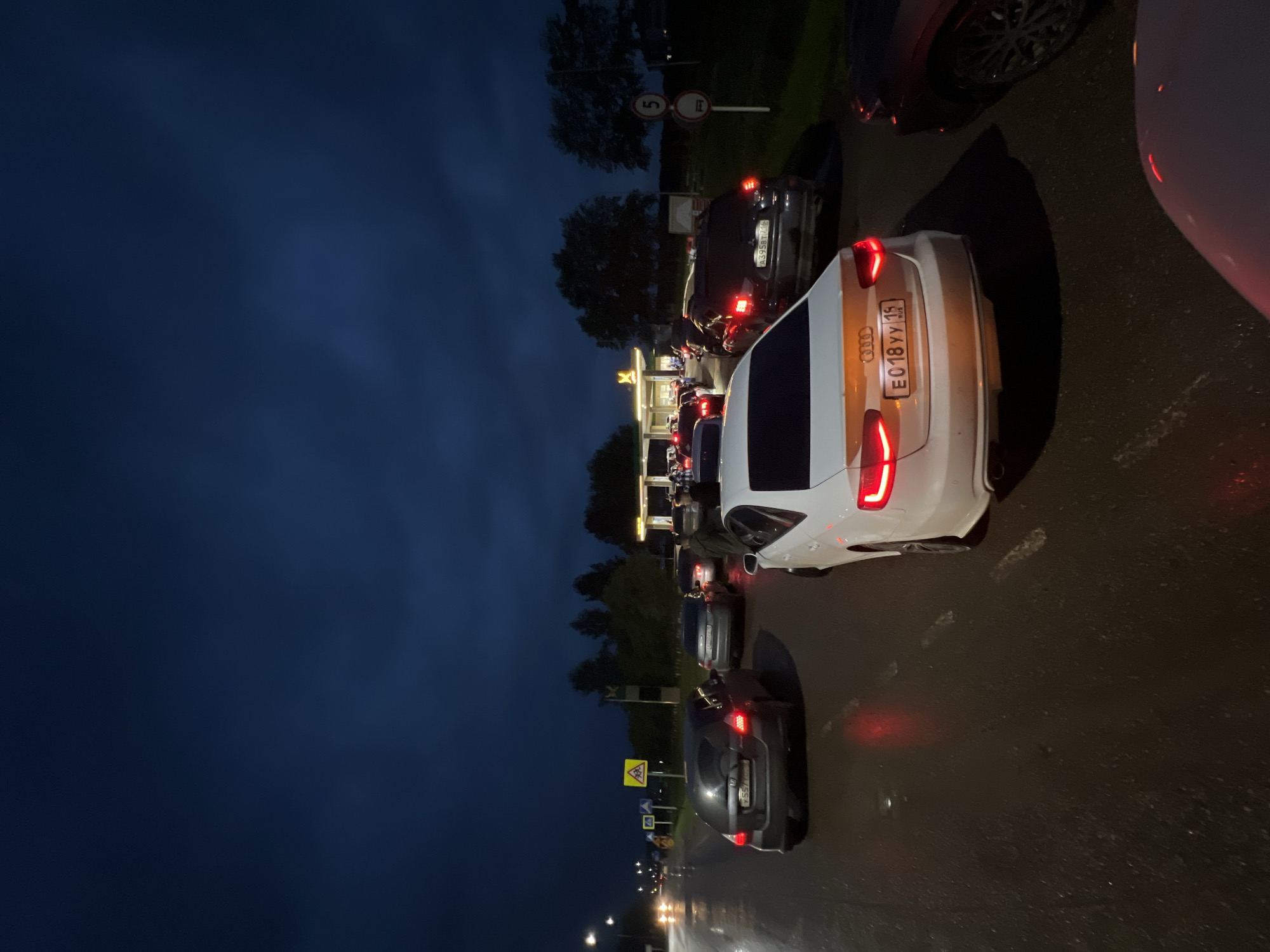 In Agryz, Russia, a traffic jam occurs at night on a wet road, likely due to a gas station. Several cars are queued up, with headlights and taillights illuminated. The sky is dark with clouds, suggesting a recent or ongoing rain. Traffic signs are visible on the side of the road, including a yellow triangular warning sign with a pedestrian crossing symbol and a circular sign with the number 5 and a prohibition symbol. The gas station, with its illuminated canopy and pumps, is the focal point of the congestion.