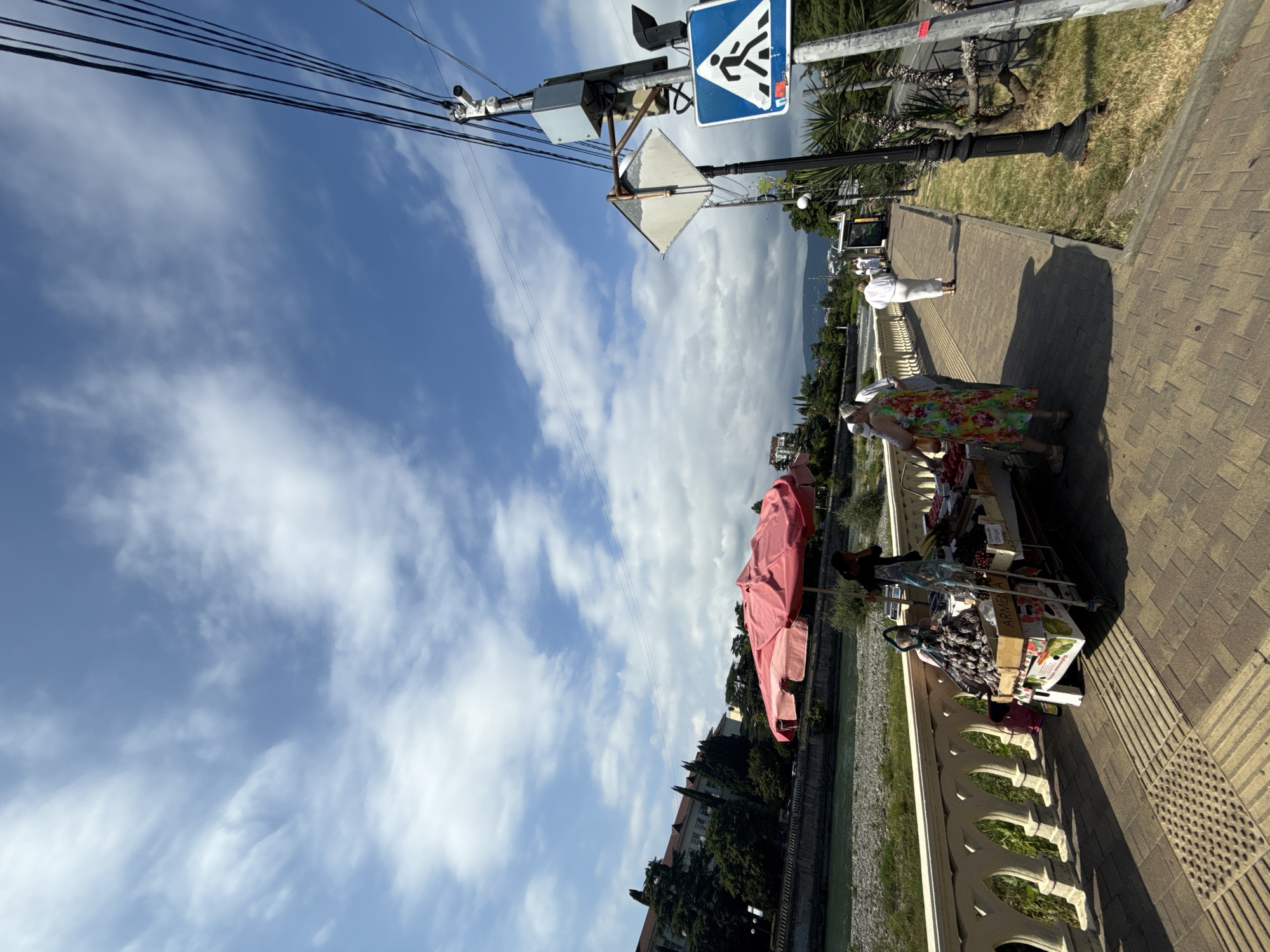 The image captures a sunny day scene in Sochi, Russia. The sky is a vibrant blue with scattered white clouds, suggesting fair weather. The foreground shows a paved walkway with a decorative railing, bordering a body of water that appears to be a canal or a river.
On the walkway, two individuals are visible. One person, wearing a floral dress, is looking down at a cart laden with goods. The cart is filled with boxes of fruits and other items, possibly for sale. Next to them, another person is partially visible. Further along the walkway, a person in white is walking away from the camera.
Several objects are present, including a street lamp, traffic signals, and signage. A pedestrian crossing sign is prominent, indicating a crosswalk nearby. Overhead, power lines are strung across the sky. A large pink umbrella is set up, casting a shadow.
The scene suggests a casual daytime activity, possibly a marketplace or a promenade where vendors display their wares. The presence of palm trees and lush greenery hints at a warm climate and a coastal or resort-like setting. The overall impression is one of everyday life on a pleasant day in a public outdoor space.