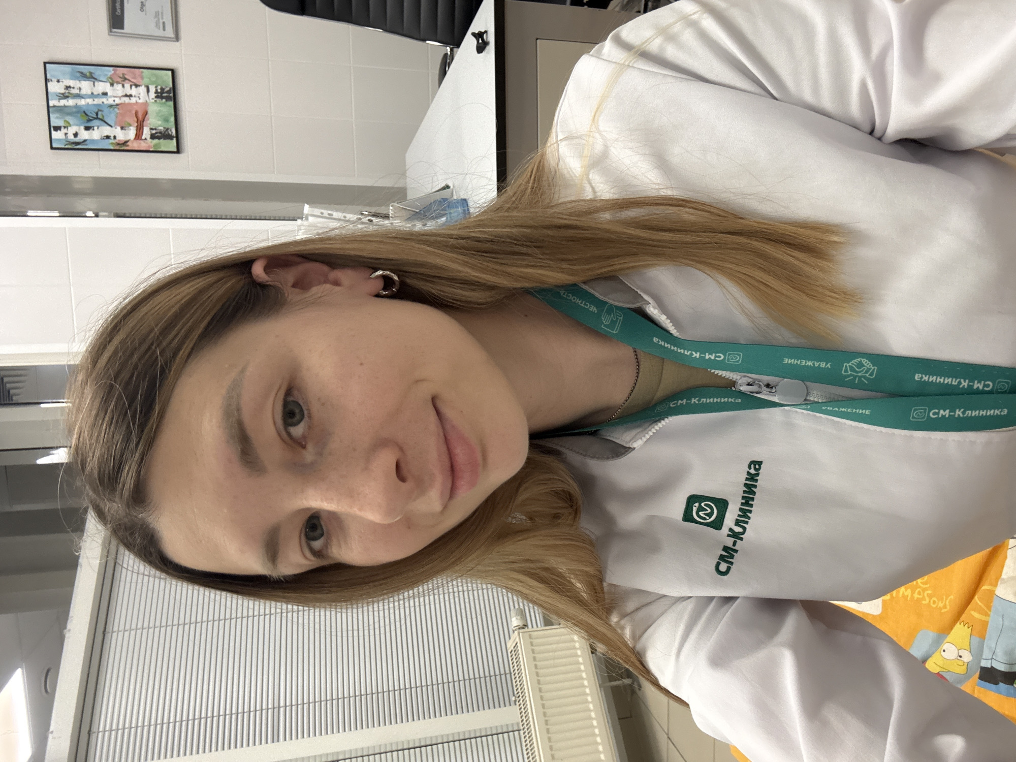 A young woman with shoulder-length blonde hair and light skin is photographed from the chest up, facing the camera and smiling faintly. She is wearing a white lab coat with a green logo and text that reads "СМ-Клиника" (SM-Klinika) in Cyrillic. A green lanyard with the same logo and text, along with the word "УВАЖЕНИЕ" (Respect), is visible around her neck. She has a small hoop earring in her visible left ear.
The background suggests an indoor, office or clinic setting. To the left, a radiator is visible beneath a window covered with horizontal blinds. The wall behind her is white and tiled. On the right wall, a small framed picture of birch trees hangs, and a framed document, possibly a certificate, is also visible. A white desk is partially in view, with a black office chair behind it. The lighting appears to be artificial, likely from overhead fixtures. The overall environment suggests a medical facility within Moscow, Russia, given the Cyrillic text and common clinic aesthetics.