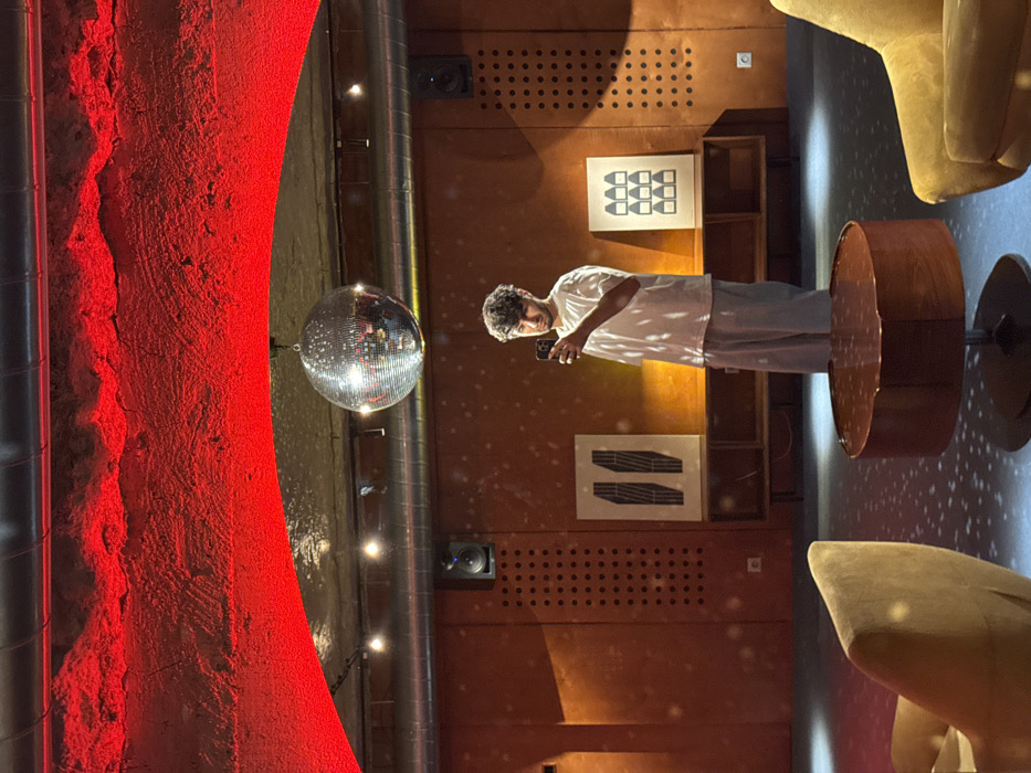 A disco ball hangs near a lit red wall. A person in a white shirt and gray pants stands in front of a wood-paneled wall with artwork, taking a photo with their phone. Beside the wall, a round wooden table sits near a yellow chair. Small spotlights line the wall near the disco ball.