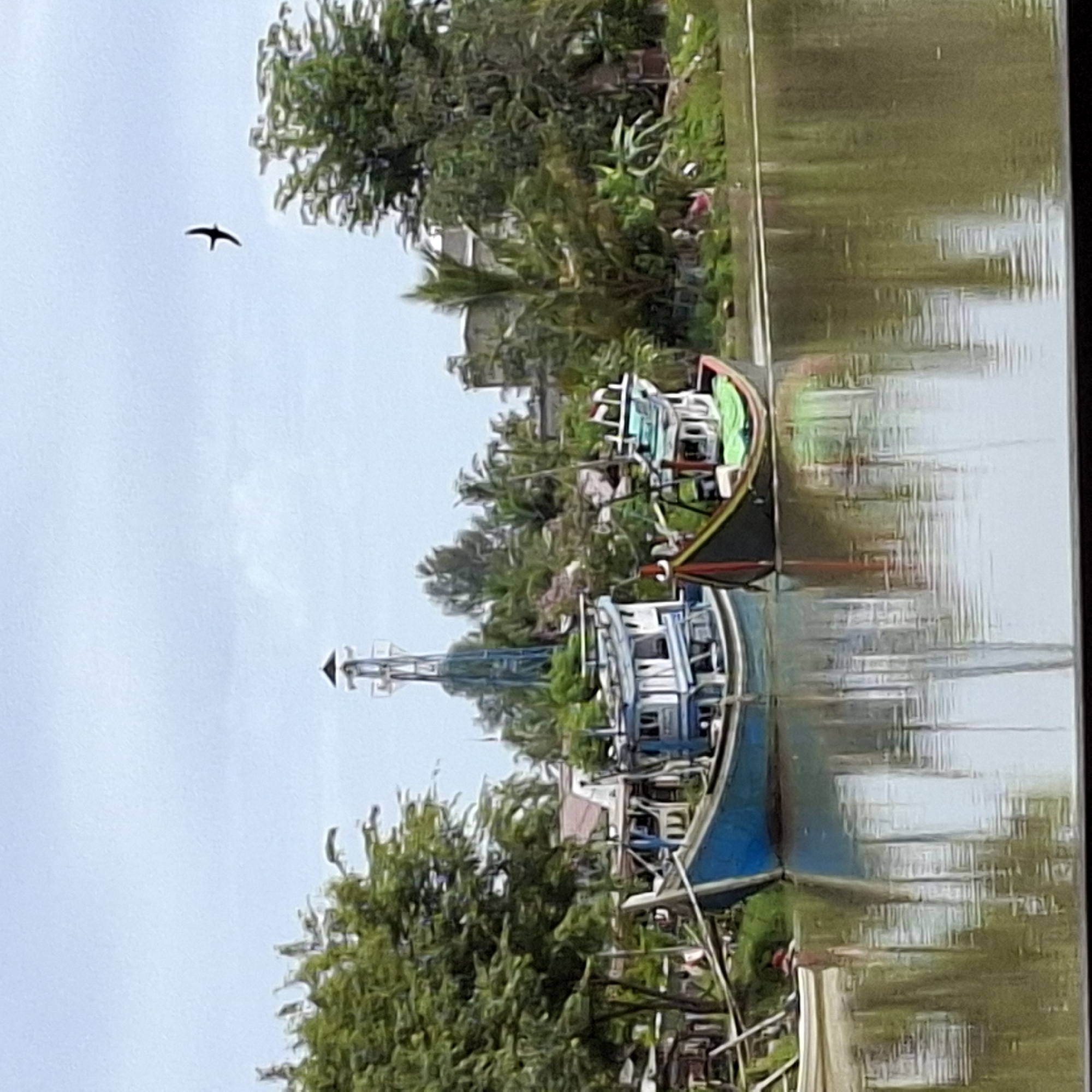 Two fishing boats are moored in a river in Mesjid Tuha, Indonesia. The boat on the left is blue and white, and the boat on the right is black and red with a green tarp. The boats are anchored near a lush green riverbank lined with trees and buildings. In the sky, a bird is soaring, and a distant lighthouse-like structure is visible behind the trees. The water reflects the sky, boats, and surrounding environment, creating a peaceful and serene scene.