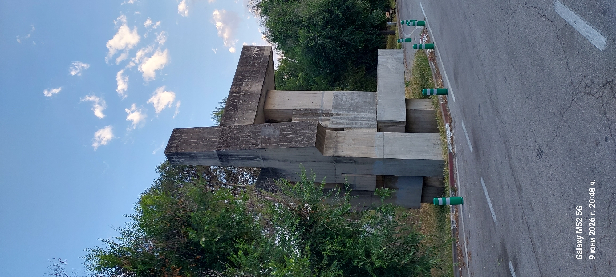 This is a full-frame, eye-level, outdoor photograph taken in Subirats, Spain. The image features a large, abstract concrete sculpture set against a backdrop of green trees and a blue sky with scattered white clouds. The sculpture is composed of geometric shapes, predominantly rectangular blocks, arranged in a complex, angular manner. In the foreground, a paved road with white lane markings is visible, with several green and white striped bollards lining the edge of the road. The overall impression is one of a monument or art installation in a public space. The timestamp overlay at the bottom left indicates the image was captured on June 9, 2026, at 20:48.