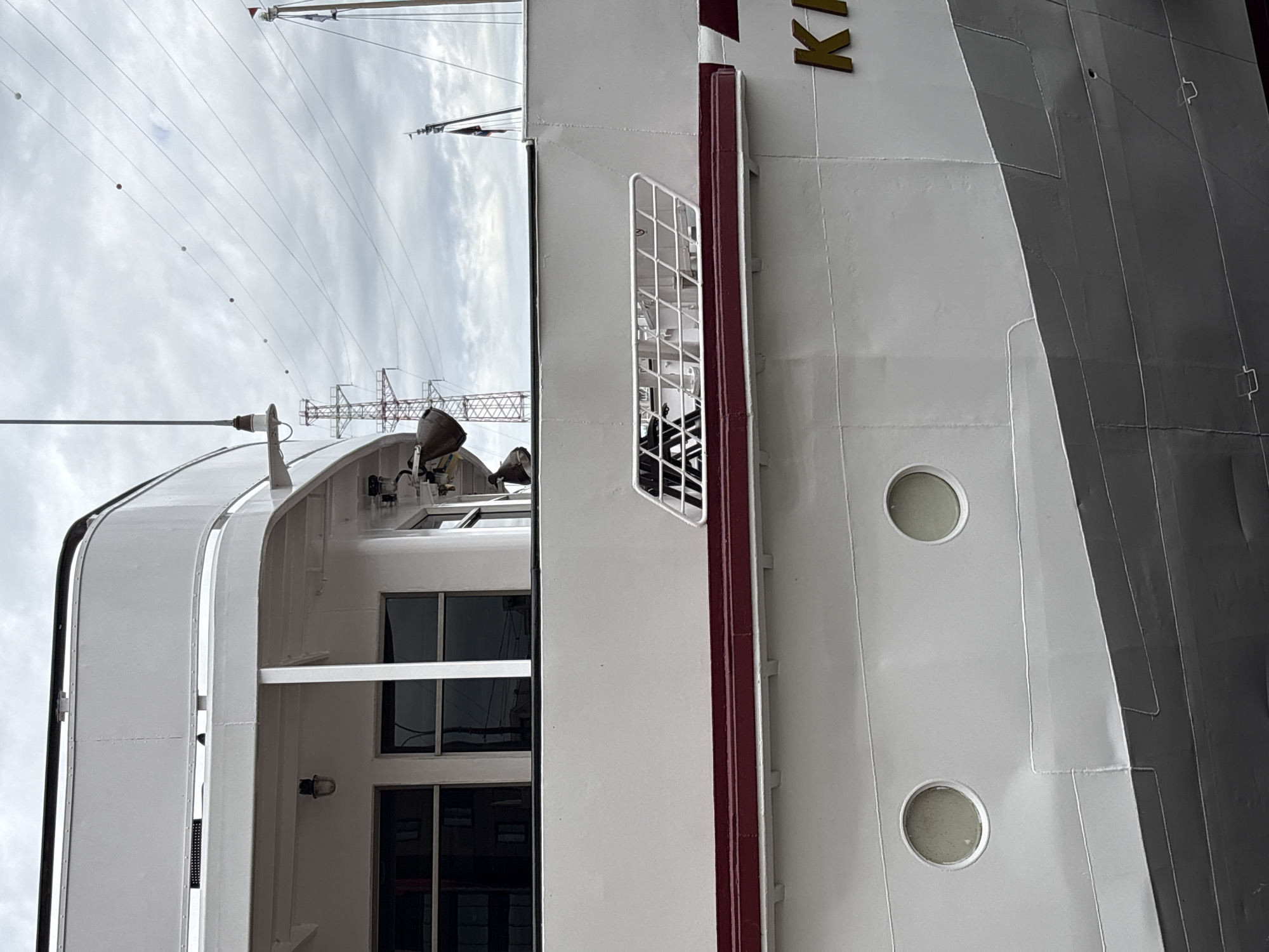 A section of a white ship's hull is visible in an outdoor, overcast setting. The hull has a dark red stripe running horizontally along its length, and above this stripe are two circular windows. Further up, there is a grated ventilation opening. The ship appears to be docked, as evidenced by the rigging and power lines visible in the background. The name "KI" is visible in gold lettering on the hull. The location is identified as Shushary, Russia.