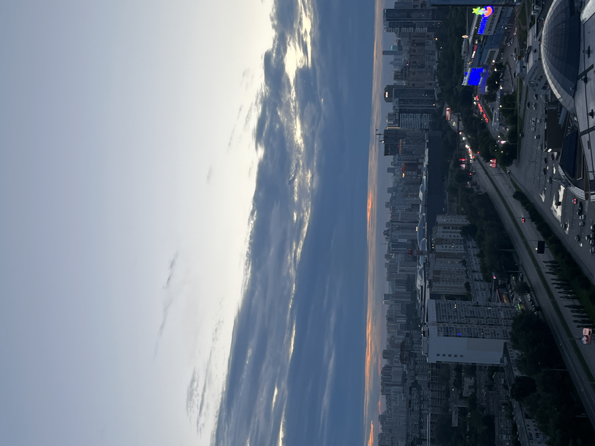 The image depicts an aerial view of Yekaterinburg, Russia, at dusk. The sky is a gradient of pale blue and grey, with elongated clouds streaked with darker blues and hints of orange and yellow from the setting sun near the horizon. A dense urban landscape stretches across the frame, characterized by a mix of residential buildings, high-rise office towers, and commercial structures.
In the foreground, a large, dark, domed building is visible, with a portion of its rooftop and surrounding parking lot occupying the bottom right corner. Cars are parked in the lot, and some are visible on a wet surface, suggesting recent rain.
A multi-lane road runs through the mid-ground, with vehicles creating streaks of light, indicating traffic flow. The road is flanked by trees and sidewalks. Adjacent to the road are several commercial buildings, one of which features a large, brightly lit blue digital display and a sign with colorful cartoon characters. Another building displays a sign with Cyrillic lettering, "АНДЕРСЕН" (Andersen).
Further into the cityscape, a variety of buildings, from older, block-style residential complexes to modern skyscrapers, fill the horizon. Some buildings have illuminated windows, indicating occupancy. A prominent white high-rise residential building stands out in the mid-left of the frame. Construction cranes are visible among the taller buildings in the distance. The overall impression is of a large, active city at the transition from day to night.