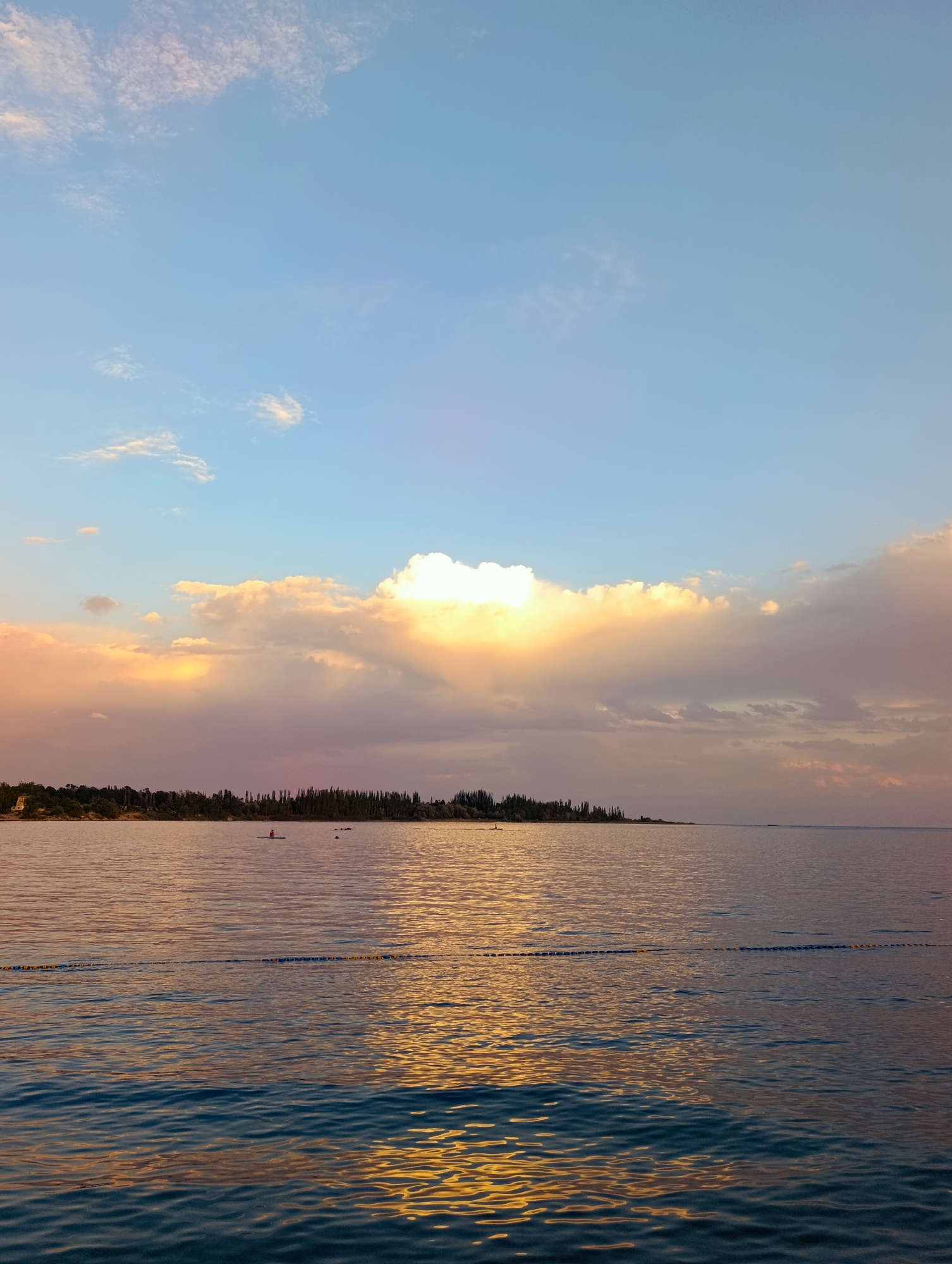This is a photograph taken at sunset over Issyk-Kul Lake in the city of Bayet, Kyrgyzstan. The sky is a gradient of blue, with large, fluffy clouds illuminated by the setting sun in shades of orange, yellow, and pink. A ray of sunlight breaks through the clouds, casting a bright spotlight on the water. The lake's surface reflects the colors of the sky, with shimmering golden and purple hues. In the distance, a shoreline with a dense line of trees is visible, appearing as a dark silhouette against the colorful sky. On the water, there are a few small figures. One person is visible on a paddleboard in the middle of the lake, and two other dark, indistinct shapes are also on the water. A yellow and blue rope barrier stretches across the water in the foreground. The overall scene depicts a tranquil and picturesque sunset over a large body of water.