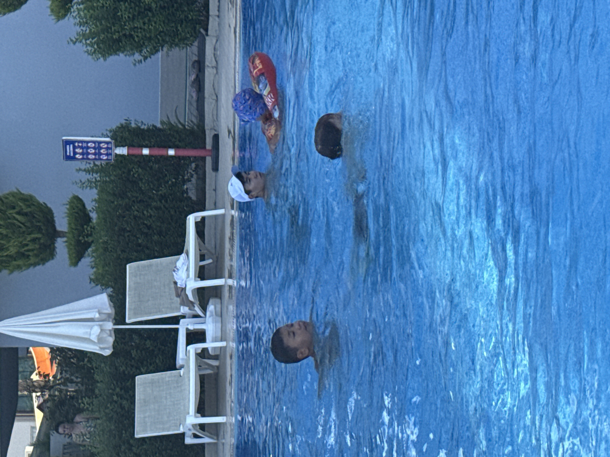 This image shows a swimming pool at the Kleopatra Beach Hotel in Alanya, Turkey. The pool is filled with blue water and there are several people swimming in it. In the background, there are lounge chairs and umbrellas. There is also a sign with pool rules. The sky is blue and there are some clouds. The hotel is a popular tourist destination in Alanya, and the pool is a great place to relax and cool off.