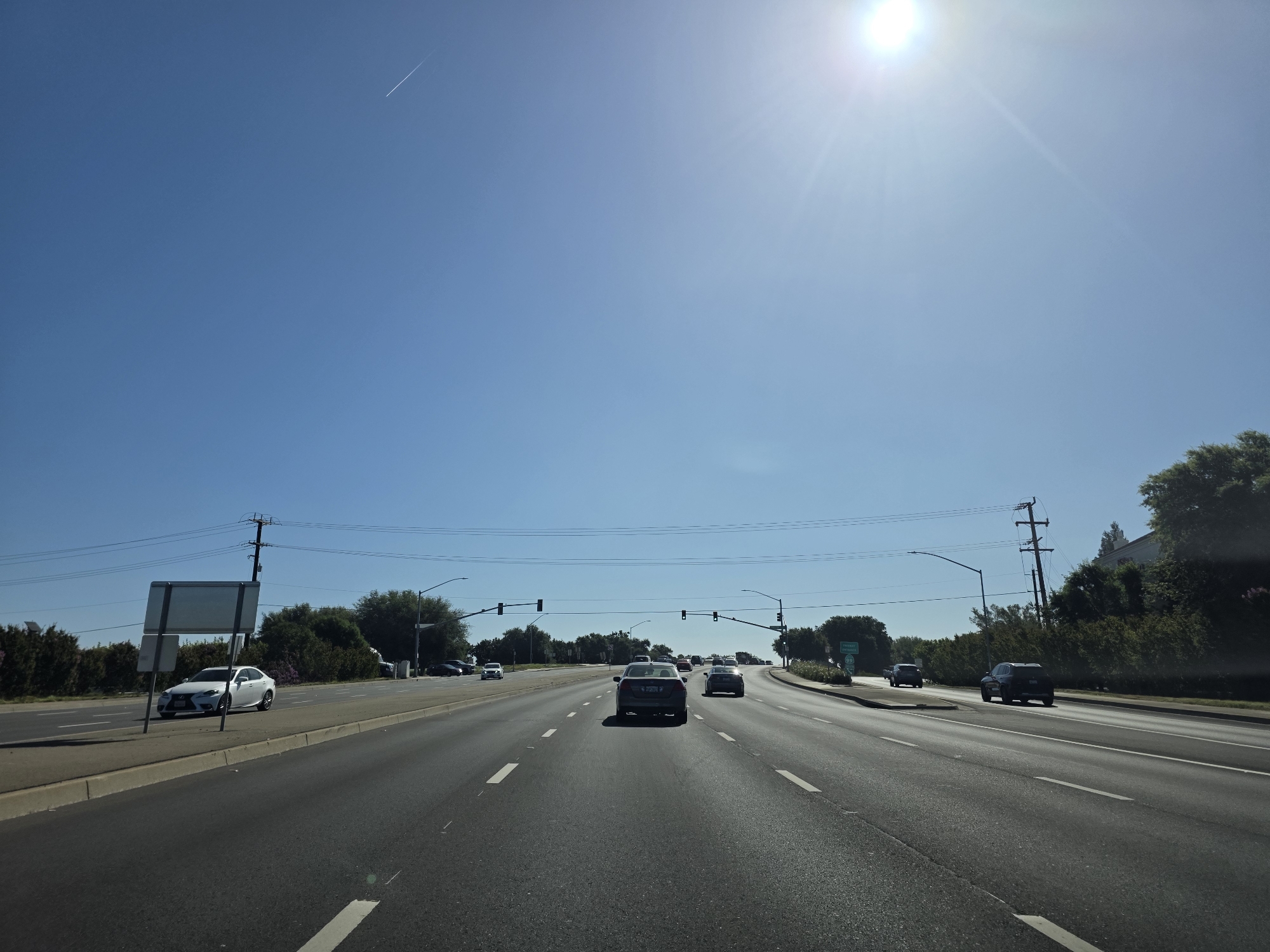 This media file captures a wide-angle view of a multi-lane road in Elk Grove, USA, under a clear, bright blue sky. The sun is high in the sky, suggesting it is midday. The weather appears to be sunny and warm.
The primary subjects are the road and the vehicles on it, indicating a scene of everyday travel. Several cars are visible, moving in the same direction as the camera. In the foreground, the asphalt road is marked with white lane lines. To the left, there is a sidewalk and a median with some vegetation and a large, blank sign. Power lines and utility poles run along the side of the road, and a dense line of trees and bushes borders the landscape. In the distance, more trees and some buildings are visible. High in the sky, a faint white contrail from an airplane stretches across the blue expanse.
There is no discernible activity or event taking place other than vehicles traveling on the road. The scene conveys a sense of ordinary daytime transit. No people are clearly visible within the vehicles.
Notable details include the bright sunlight, casting a slight glare in the upper right portion of the image, and the clear blue sky with the airplane contrail. The location is identified as Elk Grove, United States of America. There is no emotionally charged content or specific atmospheric mood conveyed.
Visible text includes a road sign on the right side of the road, partially obscured by trees, with the number "99" clearly visible, likely indicating a highway number. Another sign on the left side is blank and appears to be a directional or informational sign.