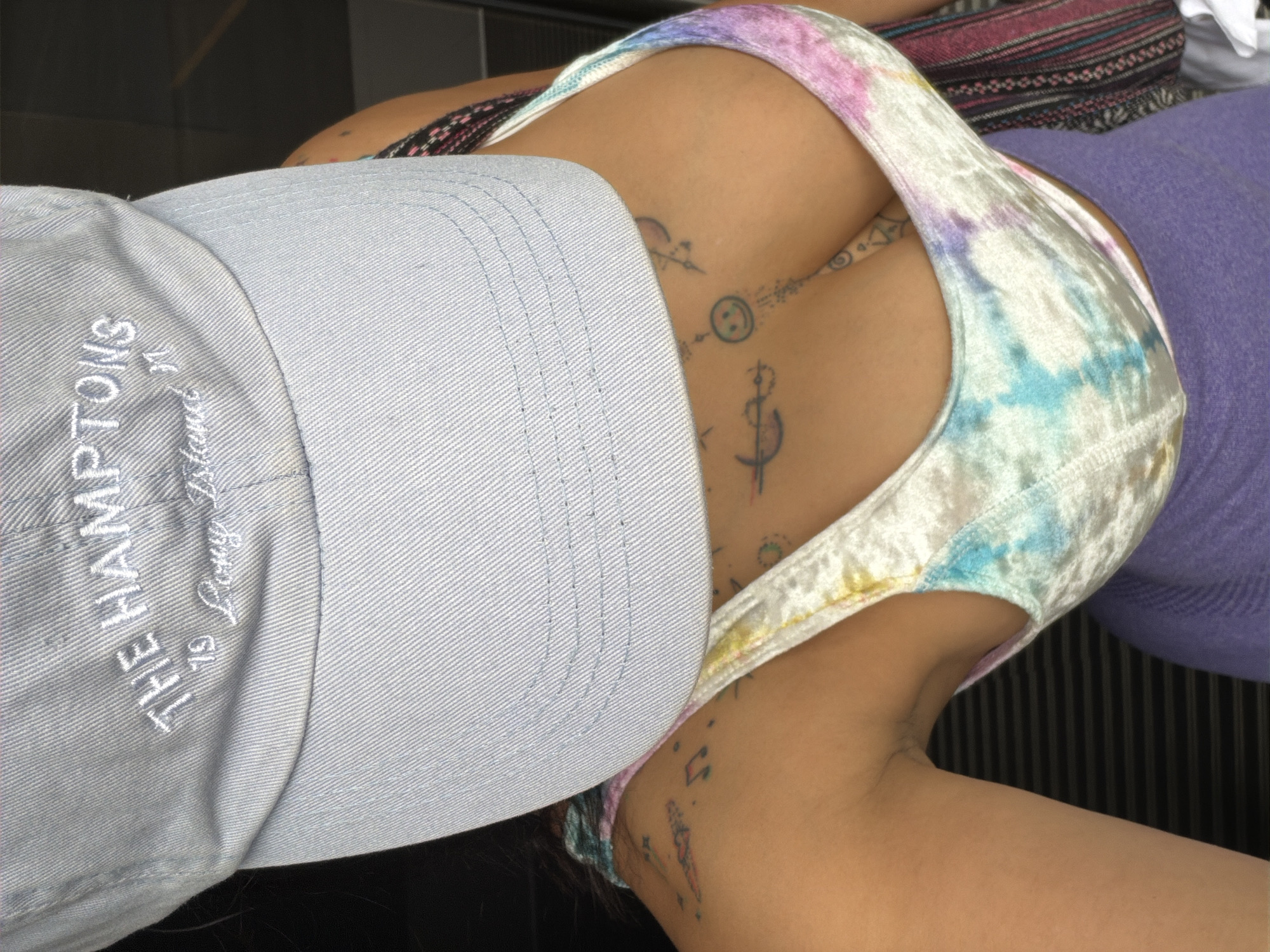 The image shows a close-up, overhead view of a person wearing a light blue baseball cap with white embroidery that reads "THE HAMPTONS 79 Long Island N". The cap is tilted down, obscuring the person's face. Below the cap, the person is wearing a tie-dye crop top with colors including white, pink, blue, and yellow. Tattoos are visible on the person's upper chest and shoulder, including musical notes and geometric designs. The background is dark and out of focus, but appears to be indoors with some horizontal lines suggesting blinds or a textured surface. There is no identifiable information within the image that places this scene in L'Hospitalet de Llobregat, Spain.