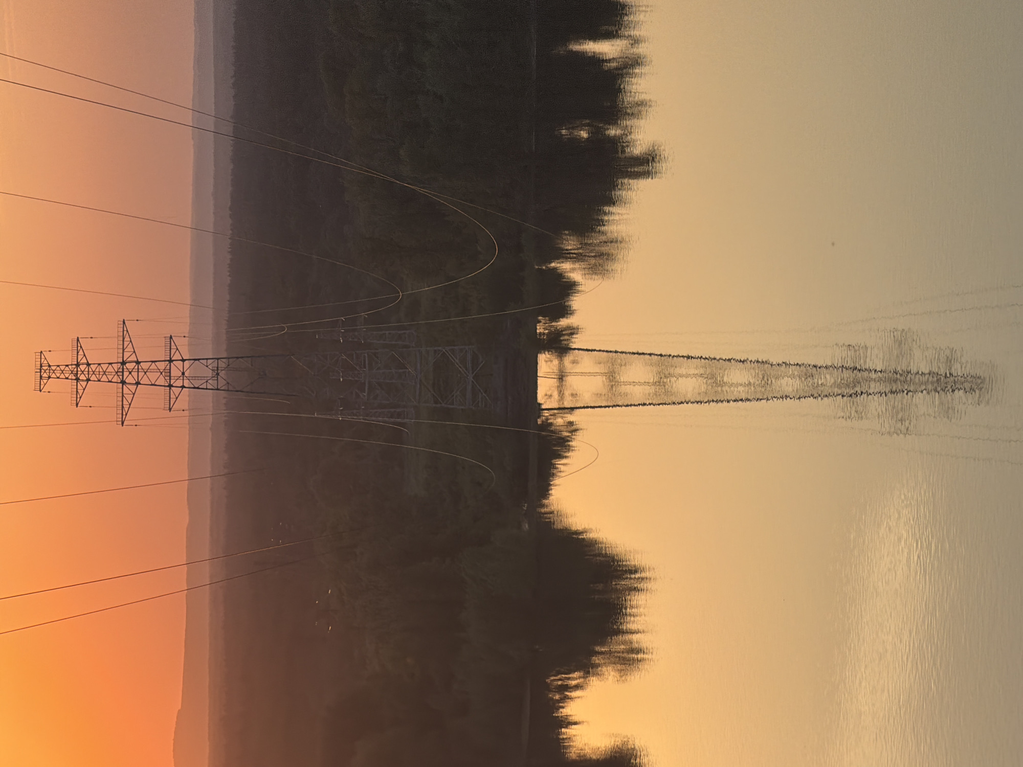 An image shows an electrical transmission tower reflected in the surface of a lake or wide river. The reflection of the tower and the lines of power transmission are vertically mirrored and extend into the water. A dark, forested shoreline is visible between the real tower and its reflection. The sky is tinted in hues of orange and yellow, suggesting a sunset or sunrise. The image appears to be taken in Noviy, Russia, based on the presence of the transmission tower in relation to the water and shoreline.