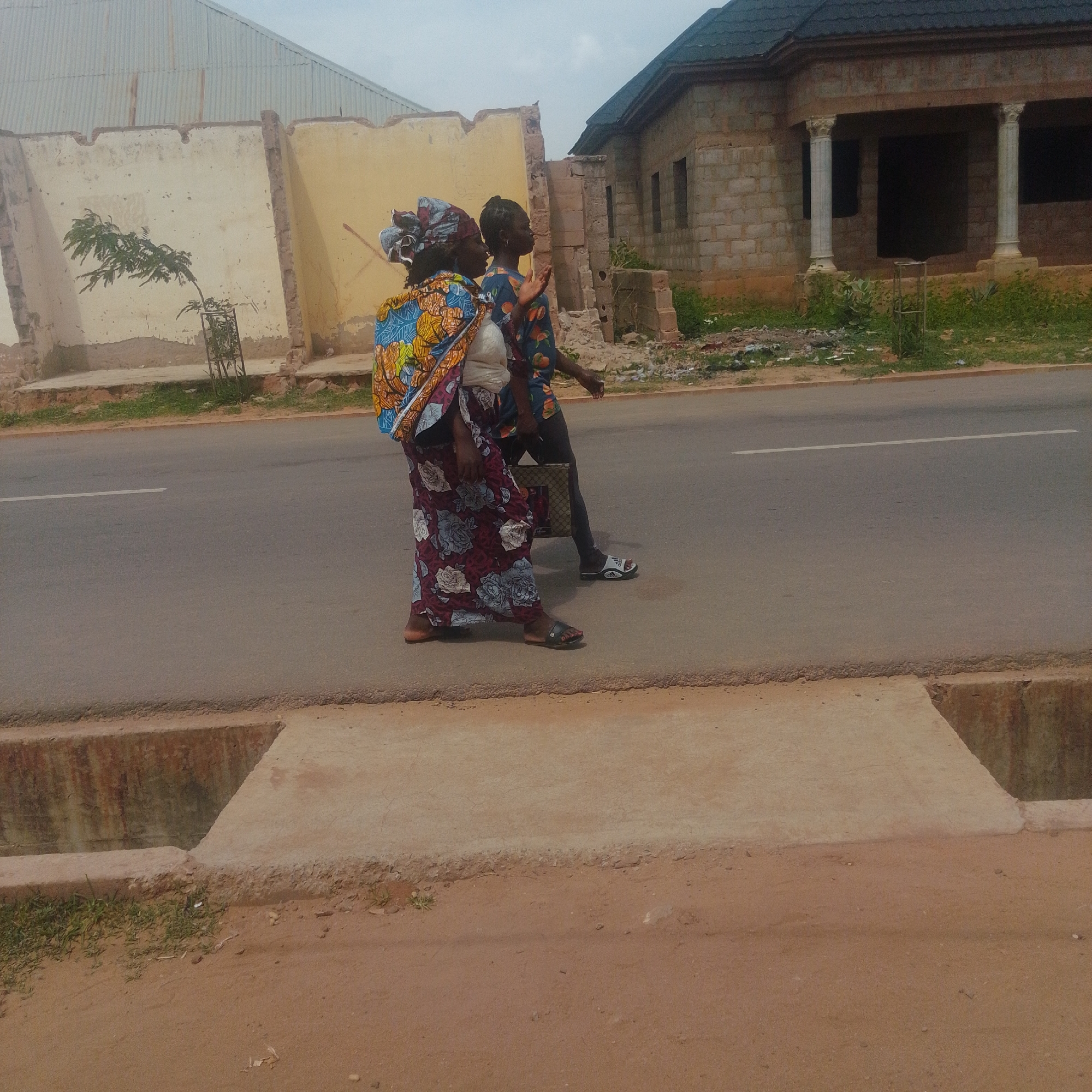 Two individuals are walking on a paved road in the city of Gombe, Nigeria. The person in the foreground, on the left, is an adult female wearing a head wrap, a brightly colored patterned wrapper, and a white top. She is carrying a baby on her back, wrapped in a blue and yellow patterned fabric. The person behind her, on the right, is an adult, possibly female, wearing a patterned short-sleeved shirt, dark leggings, and flip-flops. They are carrying a checkered bag. The road has a white line marking. To the left of the road, there is a damaged concrete wall and a corrugated metal roof of a building. To the right of the road, a building under construction is visible, made of concrete blocks and featuring columns. The ground to the side of the road is dusty and has some overgrown vegetation.