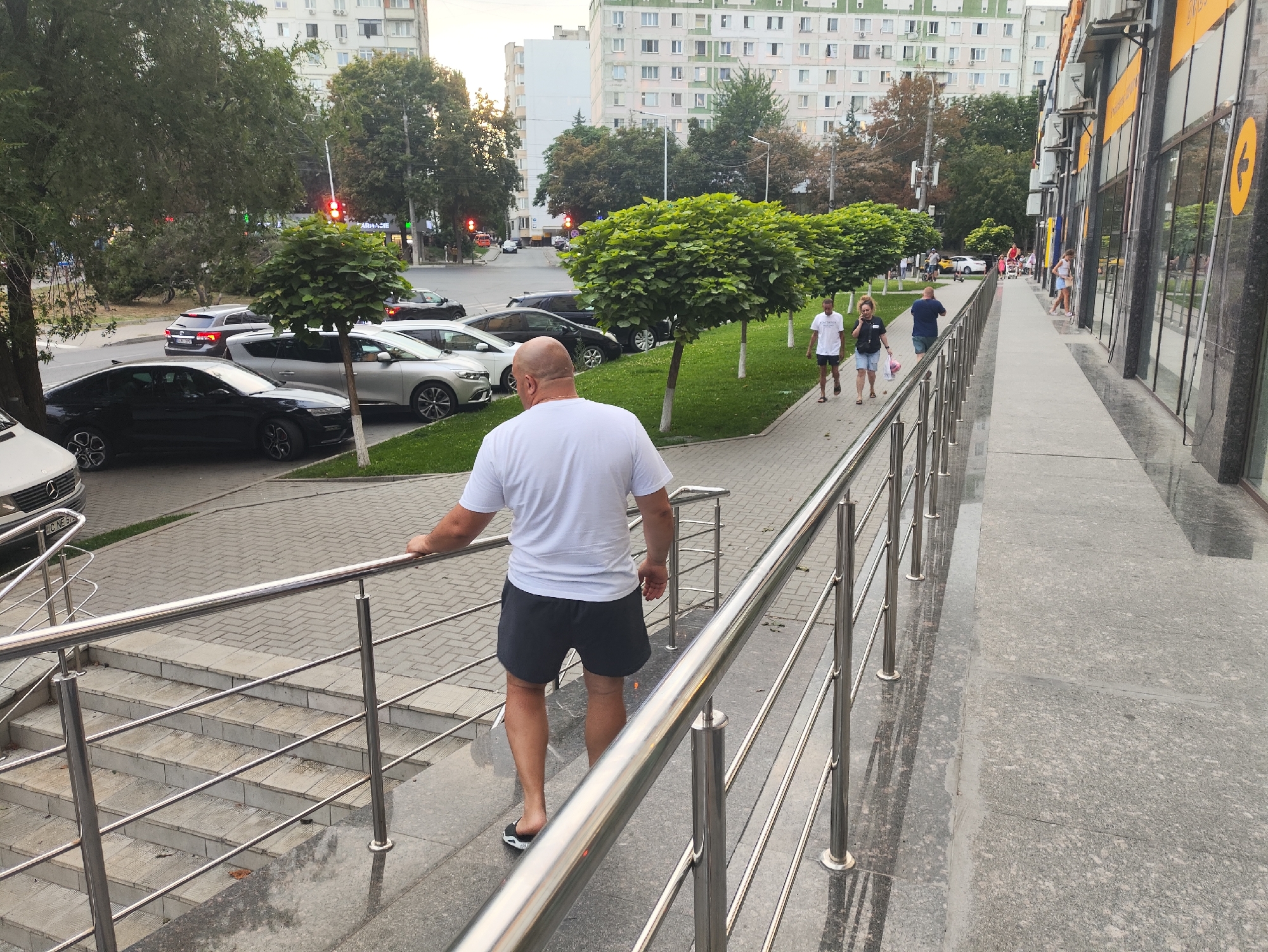 In Chișinău, Moldova, a man with a bald head, wearing a white t-shirt and dark shorts, walks up a paved outdoor staircase with metal railings. He is in the foreground, facing away from the camera. Behind him, a sidewalk with decorative trees and grass borders a street with several parked cars. In the background, apartment buildings and shops are visible. Several other people are walking on the sidewalk in the middle ground. The scene appears to be during the daytime, with overcast or late afternoon lighting.