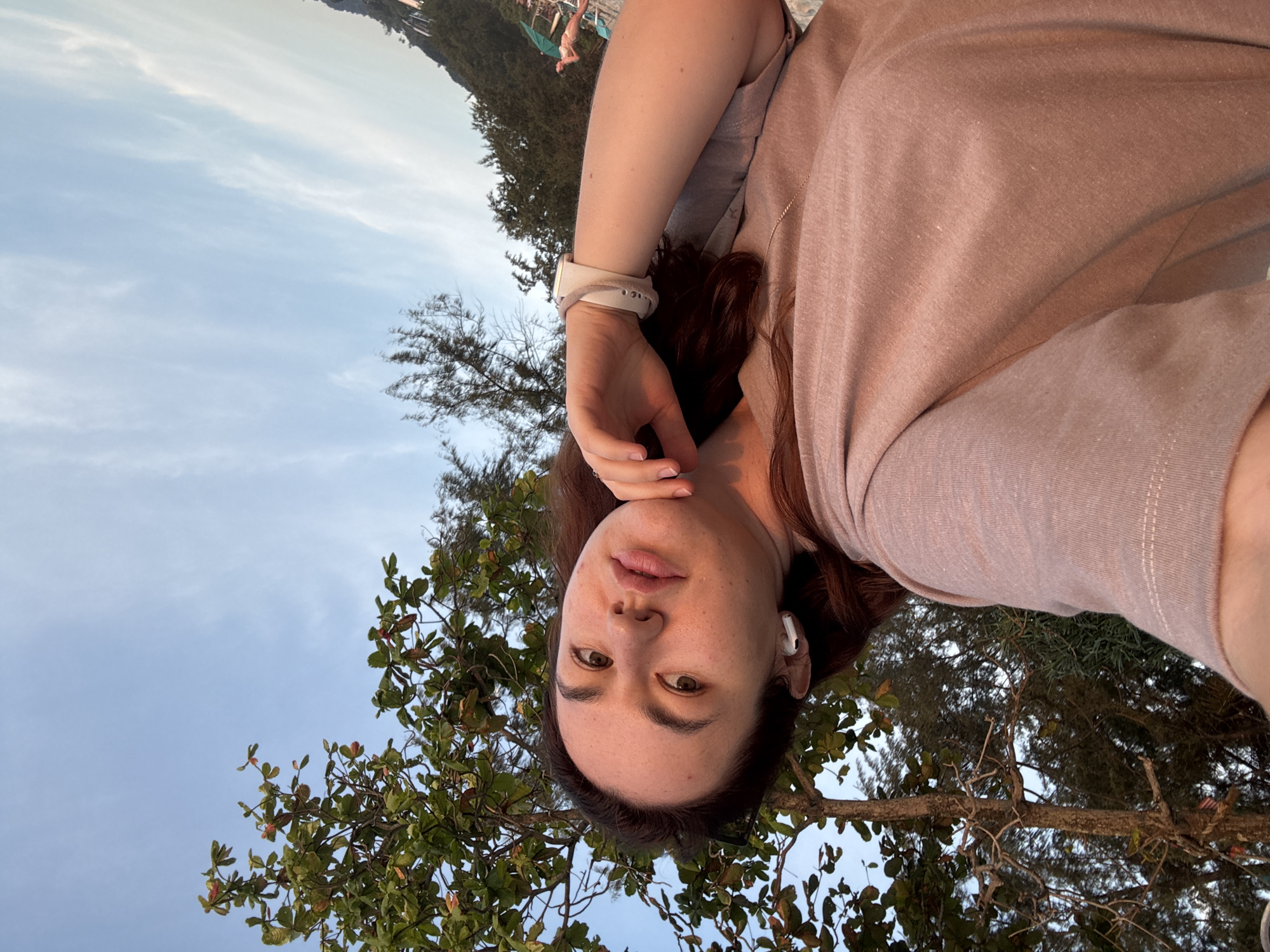 A woman is taking a selfie with her left arm extended, capturing her face and upper body. She is wearing a light pink t-shirt and white earbuds. Her right hand is resting on her chin, and she is wearing a white watch on her left wrist. Behind her, there is a tree with green leaves and a clear blue sky. In the background, a beach with people and umbrellas is visible. The image appears to be taken in Karon, Thailand, likely during sunset given the warm lighting.