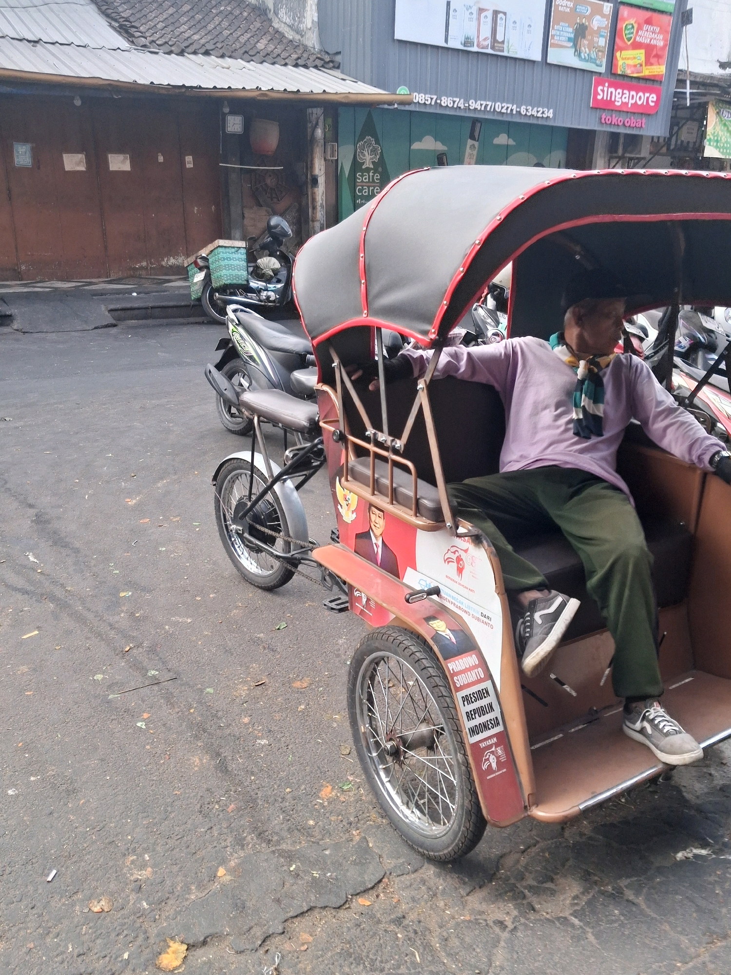 The image shows a three-wheeled motor vehicle, commonly known as a becak, parked on a paved street in Surakarta, Indonesia. A man, wearing a purple long-sleeved shirt, green pants, and a scarf, is seated in the passenger section of the becak. He is wearing dark sneakers with white laces. The becak has a canopy with red trim and is decorated with political campaign stickers featuring a portrait of a man and text in Indonesian that appears to be related to a presidential candidate. In the background, several motorcycles are parked, and buildings with brown doors and tin roofs are visible. The ground is asphalt and shows signs of wear and tear, with some debris scattered on it. The scene appears to be in an urban area during daylight.