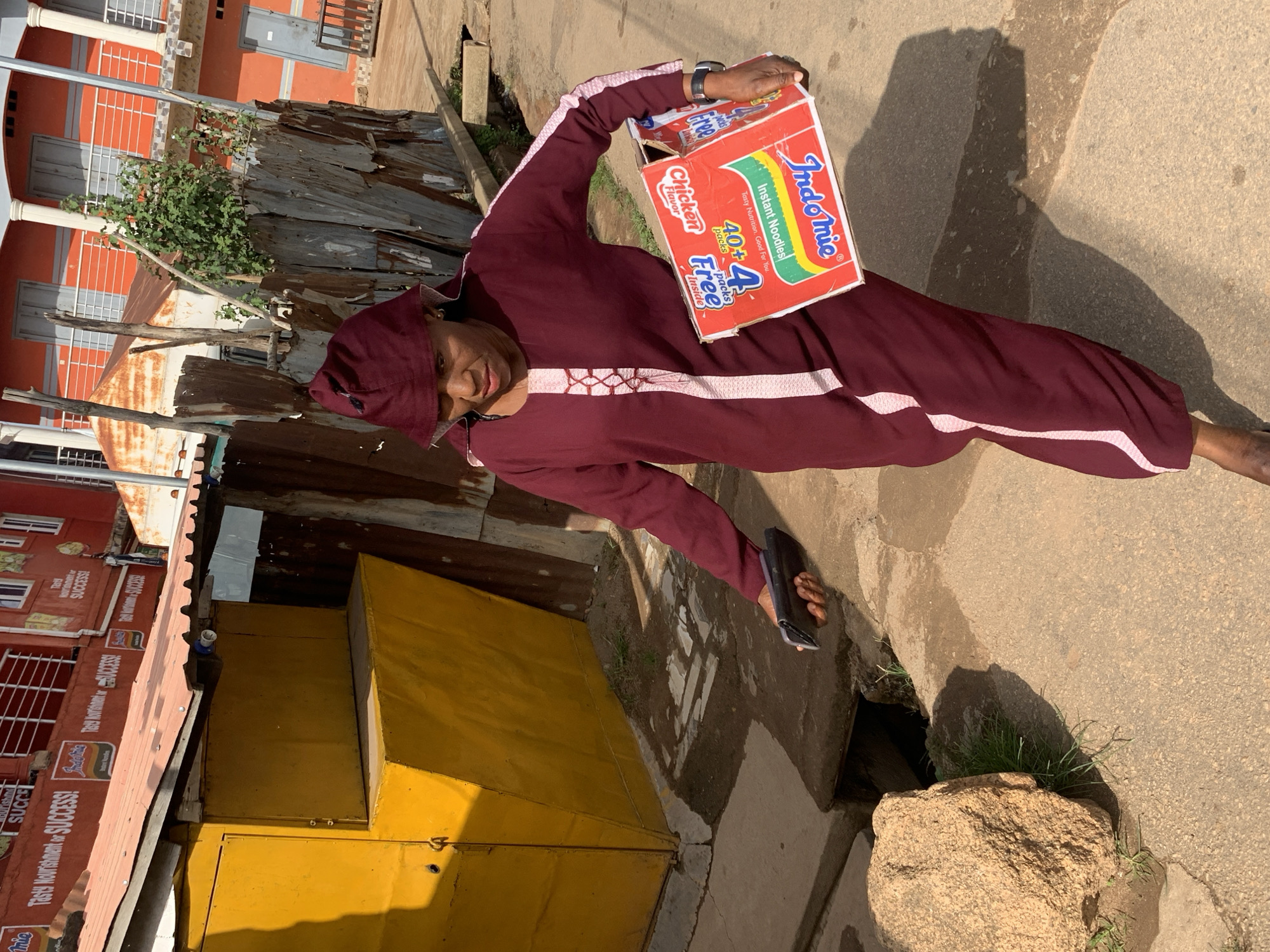 A woman walks down a sunlit street in Jos, Nigeria, carrying a red box of Indomie instant noodles. She is wearing a maroon dress and a matching head covering. She is also holding a dark-colored object, possibly a phone, in her left hand. The street is paved and appears to be a commercial area with a yellow structure and a corrugated metal fence visible in the background. The buildings in the background are painted orange and have white columns and balconies. A large rock and some overgrown grass are on the left side of the image, near a drainage channel.