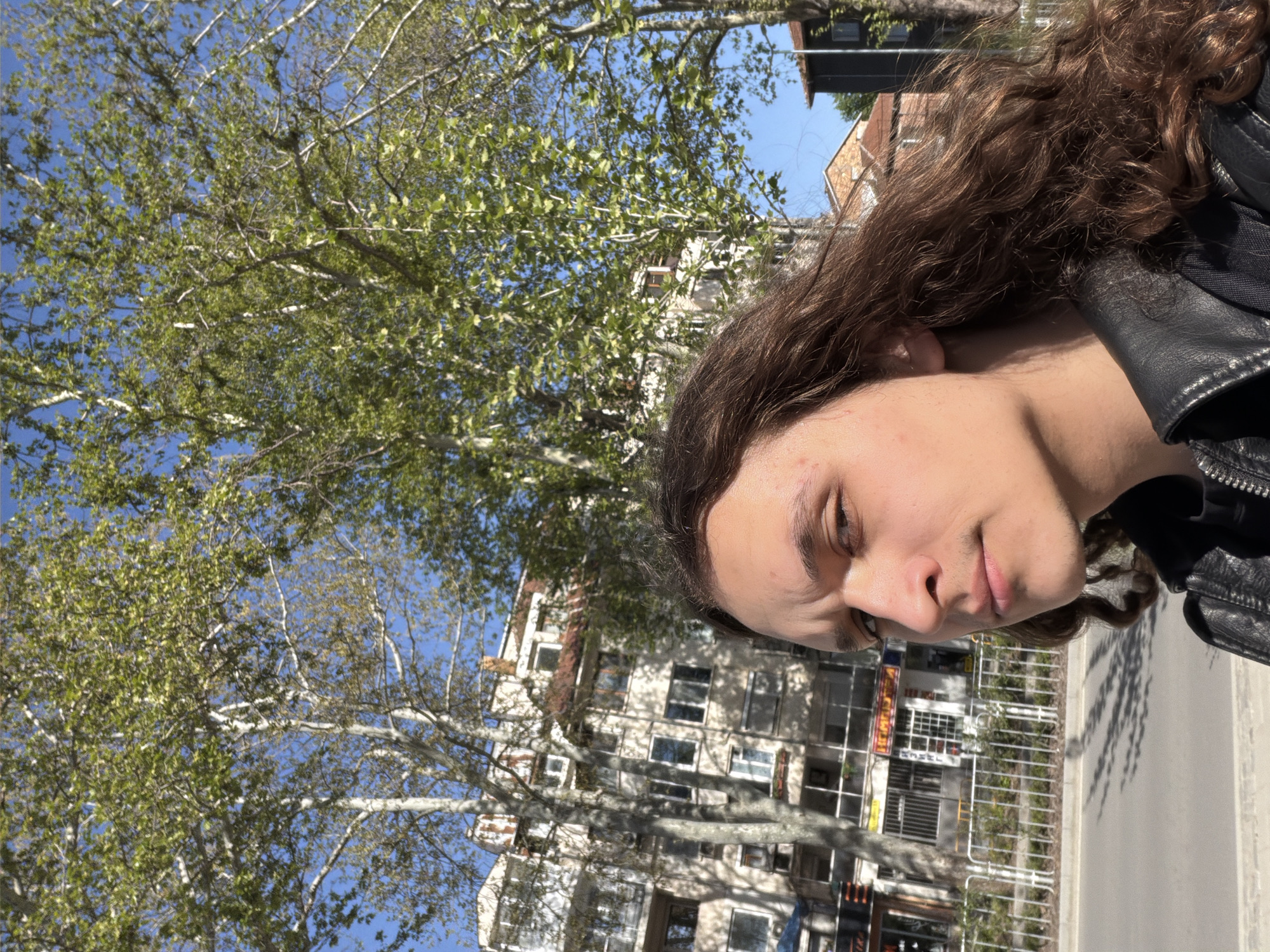 The media file is an outdoor portrait, rotated 90 degrees clockwise. When viewed correctly, it features a person in the foreground, positioned to the right of the frame, set against an urban background dominated by a large tree and buildings under a clear blue sky.
The subject is a young adult with long, wavy brown hair and light skin, wearing a black leather jacket. Their head is tilted slightly downwards, and their eyes are partially closed or looking down, giving them a relaxed or contemplative expression.
The background includes a large deciduous tree with light-colored bark and fresh, light green leaves, suggesting spring or early summer. Patches of bright blue sky are visible through the tree's canopy. Behind the tree, several multi-story buildings, likely residential or commercial structures common in urban settings, are visible, featuring numerous windows. A paved street or sidewalk with white markings and some metal railings runs horizontally across the lower part of the scene.
The image is brightly lit by natural sunlight, indicating a clear, sunny day, likely morning or afternoon, as evidenced by the distinct shadows cast by the tree. The setting is consistent with an urban street in Plovdiv, Bulgaria. No clearly legible text is visible in the image.