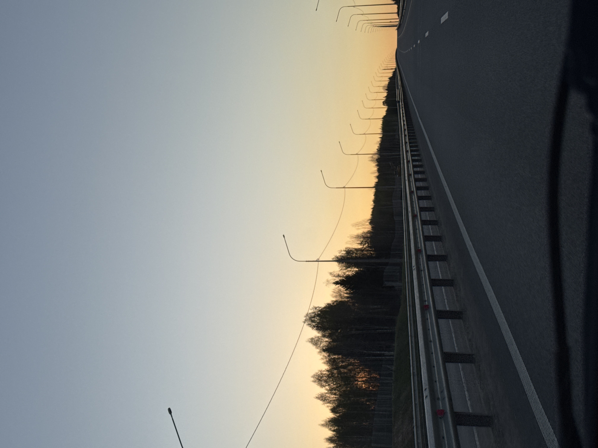 The image shows a highway in Ivanovka, Russia at dusk or dawn. The road is asphalt and has white lane markings. A metal guardrail runs along the side of the road, separating it from a line of trees. Streetlights are evenly spaced along the road, extending into the distance. The sky transitions from blue to orange as the light fades. The image appears to be taken from a moving vehicle.