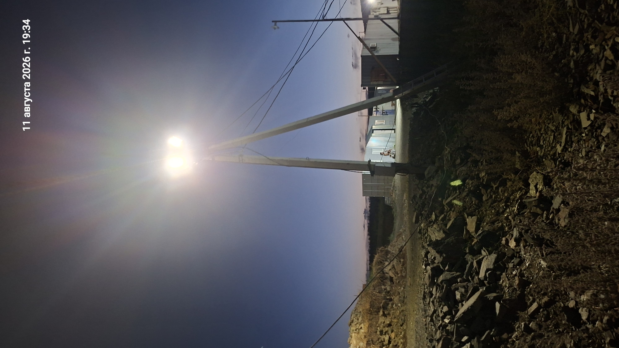 A tall, multi-panel floodlight stands on a utility pole in the foreground, illuminating a rocky, unpaved area. In the background, several modular buildings, possibly temporary structures or shipping containers, are visible. A person is seen standing near the entrance of one of the buildings. The sky is dark blue, indicating dusk or nighttime, with a date and time stamp of "11 августа 2026 г. 19:34" overlayed in the upper right corner. The scene appears to be an outdoor industrial or construction site. The mention of Maraliha, Kazakhstan, suggests this is the geographical context.