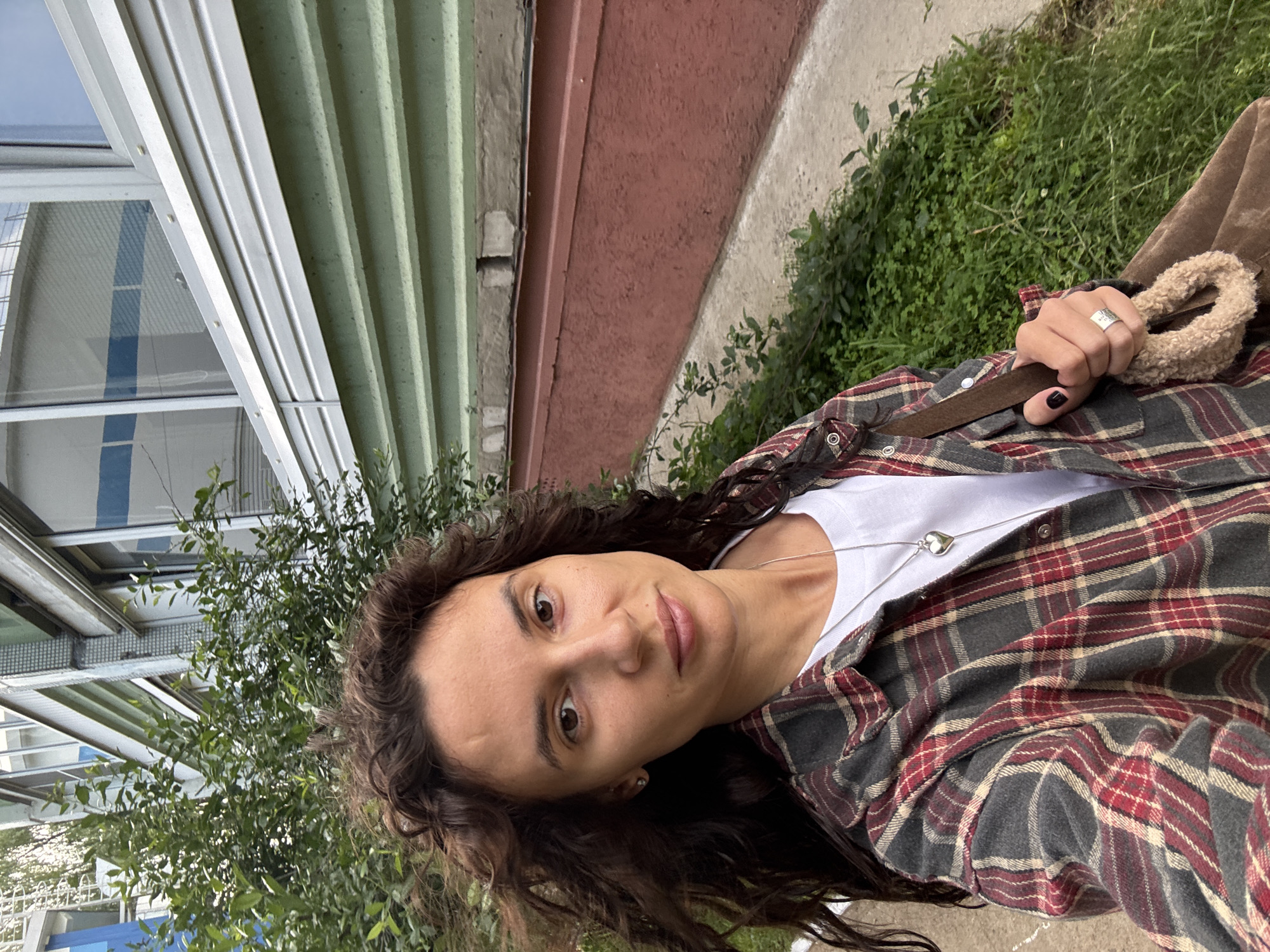 A casual outdoor portrait features a woman with long, curly dark hair, looking directly at the viewer with a neutral expression. The image is rotated, presenting her at an angle. She wears a white t-shirt beneath an open plaid flannel shirt, which has patterns in shades of grey, red, and cream. A silver necklace with a heart-shaped pendant is visible against her white t-shirt. Her right hand, featuring dark nail polish, holds a brown strap of a bag that has a fluffy, light-colored handle detail. A wide silver ring with unidentifiable engraved text is on one of her fingers.
The background consists of multiple architectural elements and natural foliage. To the viewer's left, a building with light-colored corrugated siding and a window reflecting a blue sky and horizontal white lines is visible. This transitions to a light green corrugated wall, followed by a section of red-painted concrete, and then a lighter concrete or stucco wall. Lush green foliage, including bushes and ground cover, fills the lower and right portions of the background. The scene suggests an outdoor urban or suburban environment, potentially in Chaikovsky, Russia, under soft, diffused daylight.