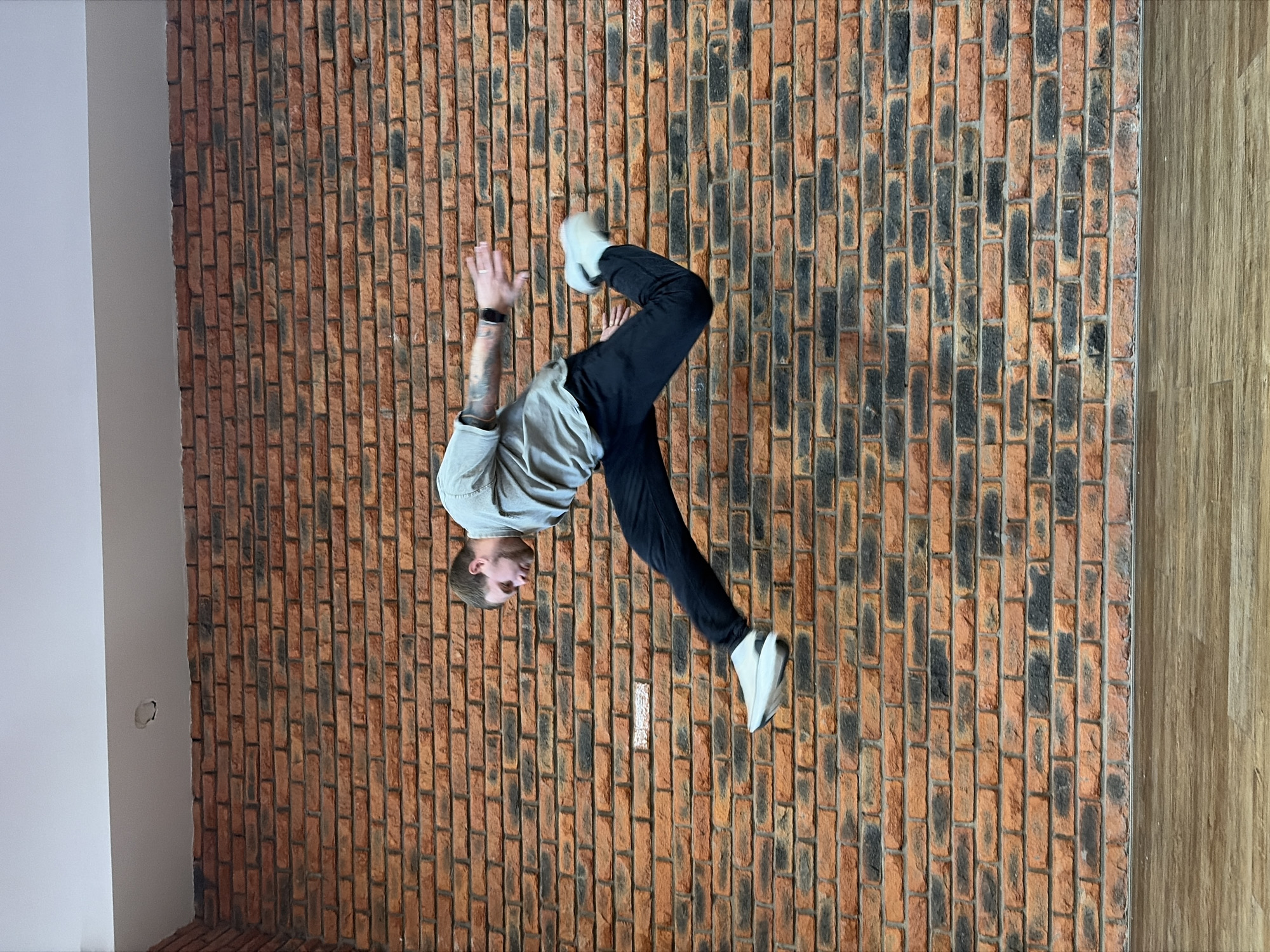 An inverted full-shot captures a young man performing an acrobatic pose, possibly a handstand or a partial flip, against a textured brick wall in an indoor setting in Chaikovsky, Russia. The man, with short hair, wears a light grey t-shirt, dark trousers, and light-colored sneakers. His body is angled, with his head positioned towards the bottom of the frame, and his legs bent and extended upwards. He has tattoos visible on his right arm and wears a dark wristband or watch on his left wrist.
The prominent background is a rustic brick wall, featuring reddish-brown bricks interspersed with darker, possibly charred or discolored, blackish sections, laid in a vertical stretcher bond pattern. To the left side of the brick wall, a plain, light grey or white wall is visible, showing a small circular imperfection or hole. To the right, a section of light-colored wooden paneling or flooring appears, indicating another distinct surface within the room. The lighting is even, suggesting an indoor environment without direct harsh sunlight. No discernible text is visible in the image.