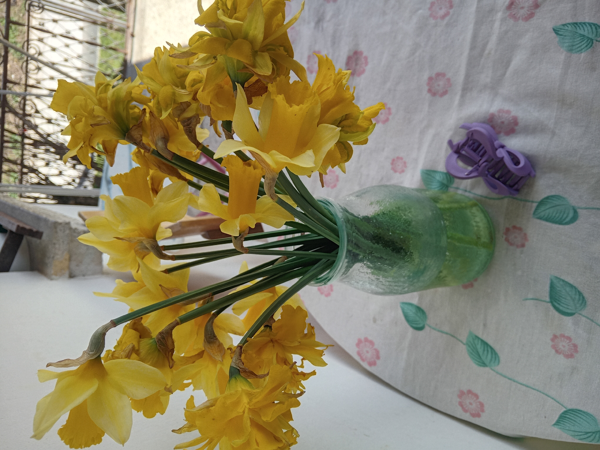 A vase of yellow daffodils sits on a patterned tablecloth, indoors in an unknown location within the city of Unknown, Unknown. A purple hair clip rests next to the vase. A window with a decorative metal grate is visible in the background. A white surface, possibly a ledge or tabletop, is beneath the tablecloth and vase.