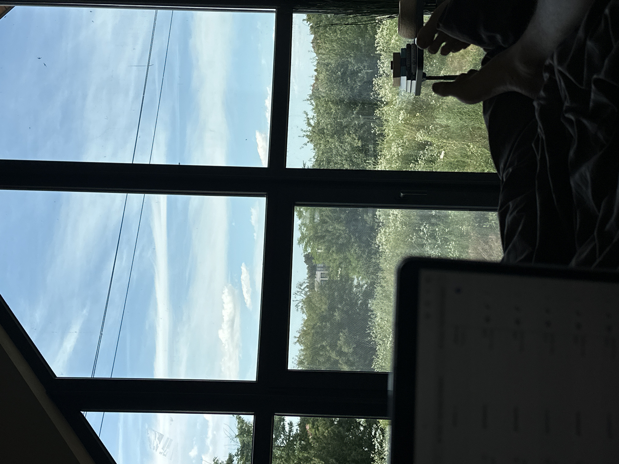 The image shows a person's hands on a laptop. The laptop screen is partially visible in the foreground, displaying a dark interface with some text and shapes. The hands appear to be actively engaged with the laptop, possibly typing or interacting with the screen. The background, visible through a large window, depicts a bright, sunny day with a clear blue sky and scattered white clouds. Outside the window, there is lush green foliage, suggesting an outdoor or nature-adjacent setting. The overall scene appears to be indoors, with the window offering a view of the outside world. The lighting suggests it's daytime, likely mid-morning or afternoon due to the brightness and clear sky. The person's hands are silhouetted against the lighter background. A stack of books or objects is visible on a stand to the right of the laptop. The scene evokes a sense of focus and possibly work or study.