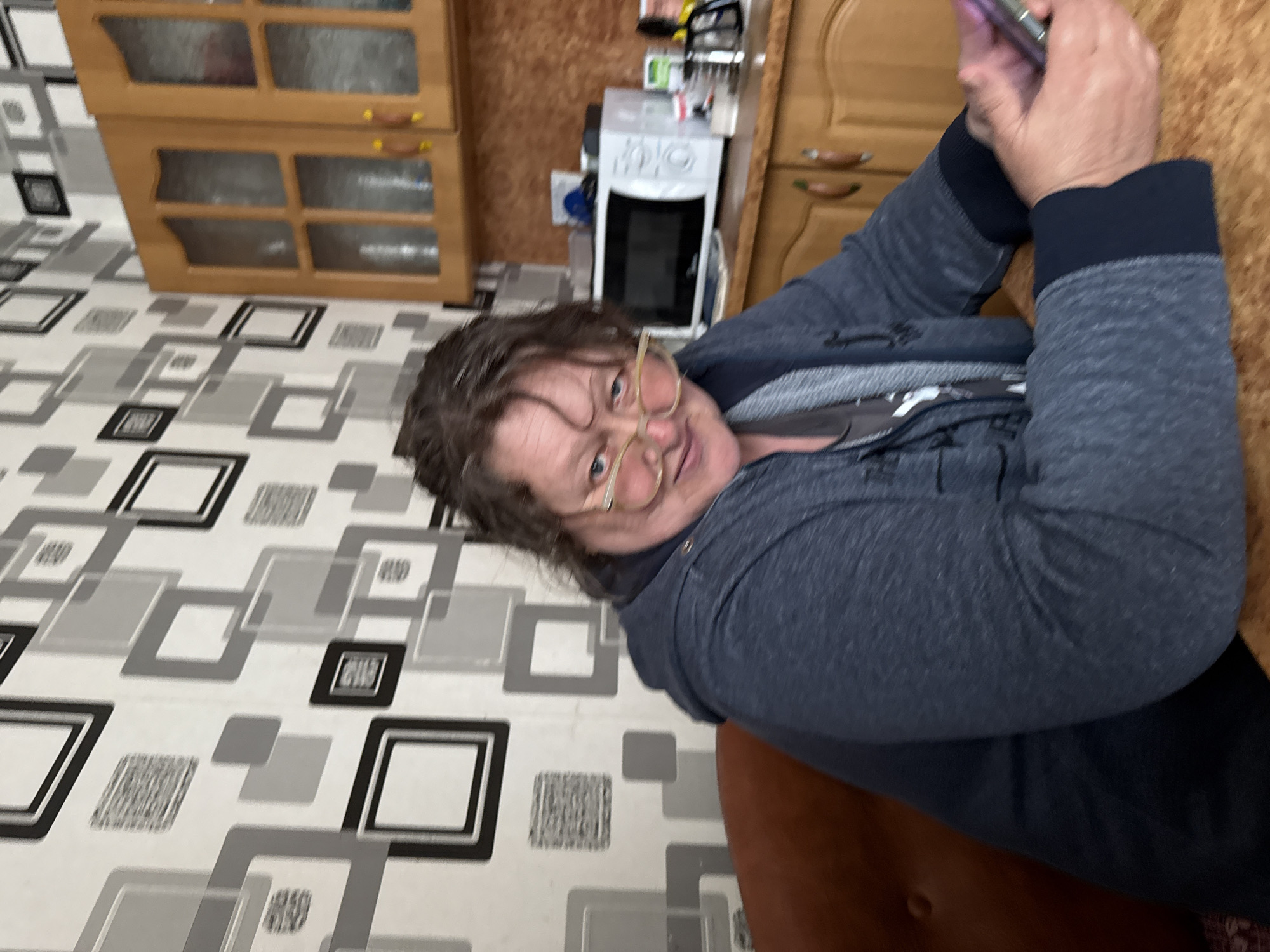 An adult woman with glasses is holding a phone and taking a selfie indoors, in Лабжанскі сельскі Савет, Belarus. She is wearing a dark grey hooded sweatshirt and sitting on a brown chair. The background includes a geometric-patterned floor, light brown wooden cabinets, and a white appliance.