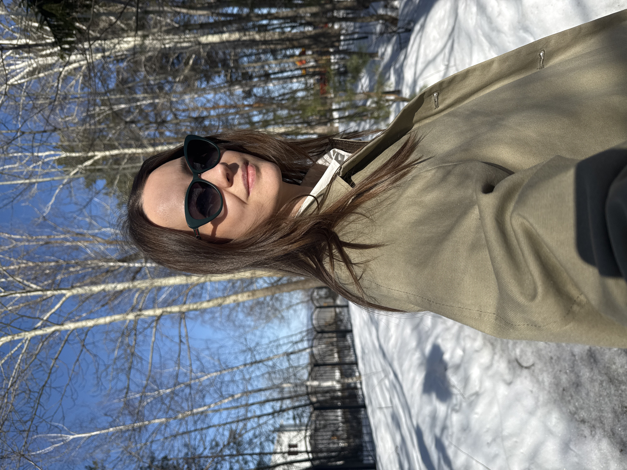 A young woman wearing sunglasses and a tan jacket is photographed in an outdoor setting, possibly Chaikovsky, Russia, based on the presence of birch trees. The ground is covered with snow. The woman faces the camera with a slight smile. The background consists of trees and a partly visible structure with fencing. The sun appears to be shining brightly, creating strong shadows.