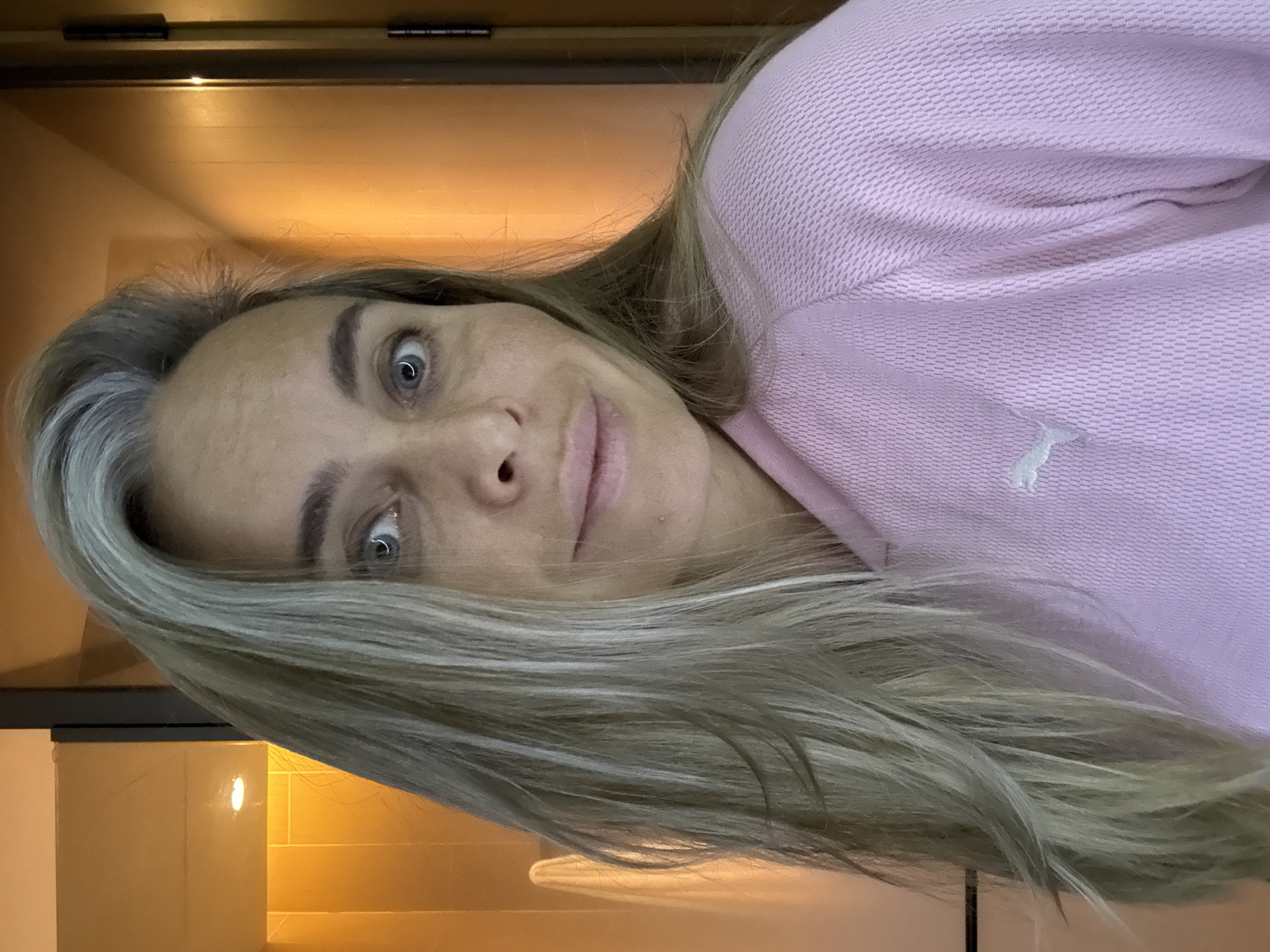 A woman with blonde and gray hair, wearing a pink Puma sweatshirt, looks directly at the camera. The background is a bathroom with orange tiled walls and a glass door. The lighting suggests it is indoors. The location is not specified as Shanghai, China, or any other city.