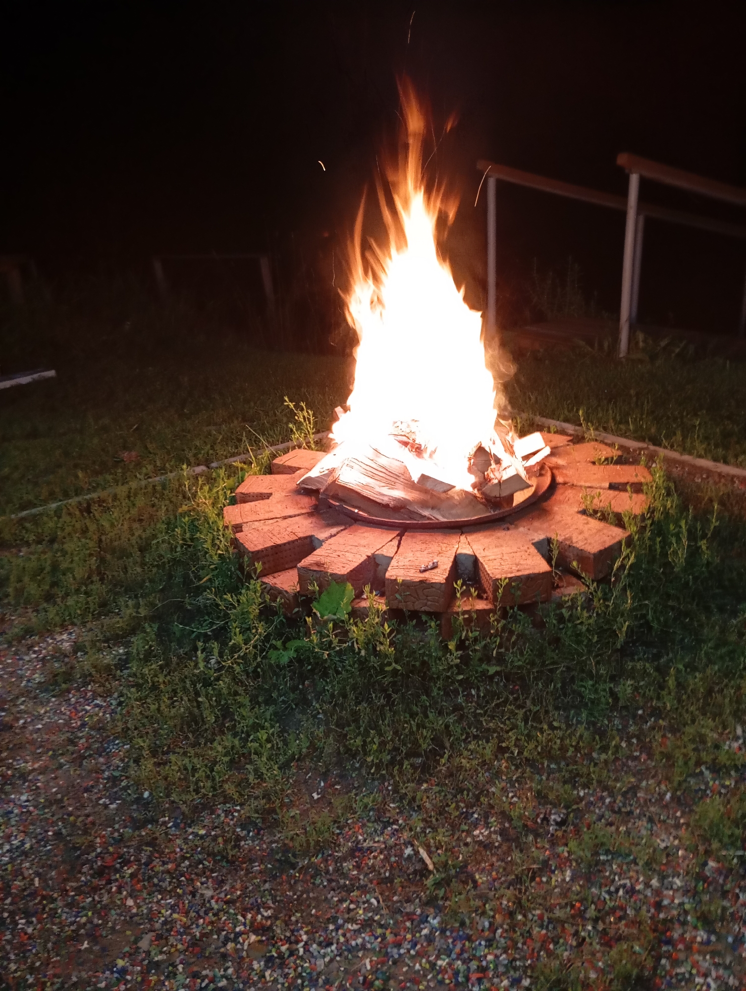 In the city of Malyi Bukor, Russia, a roaring campfire burns brightly at night, illuminating the surrounding area with a warm glow. The fire is contained within a circular fire pit constructed from bricks arranged in a sunburst pattern. Flames erupt from a collection of logs and kindling. The ground around the fire pit is covered in lush green grass, with patches of gravel and small colorful stones in the foreground. In the background, wooden railings suggest a seating area or pathway. The scene is set in an outdoor environment, likely a backyard or recreational area, during the dark hours of the night.