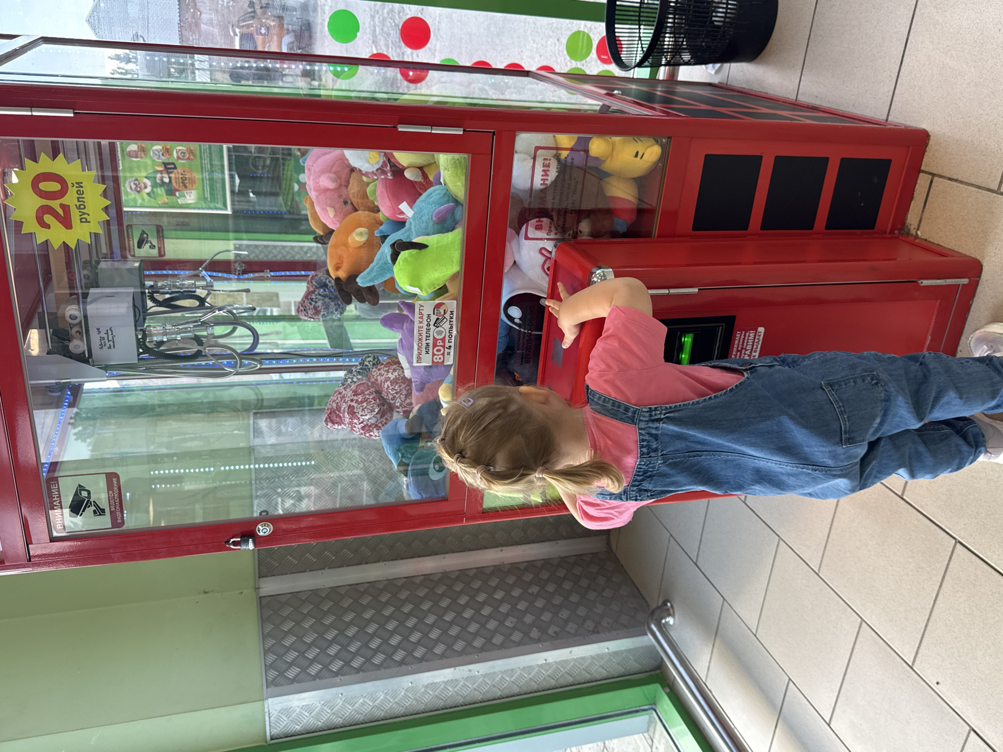 This vertical, outdoor, medium shot captures a young girl playing with a claw machine filled with plush toys. The girl, wearing a pink shirt and denim overalls, is reaching towards the machine's controls. The claw machine is bright red and has a glass front displaying various colorful stuffed animals. A sign on the machine indicates that a game costs 20 rubles. There are also signs in Russian, with one warning that video surveillance is in progress. The setting appears to be a public area with tiled flooring and a metal railing. The lighting suggests it's daytime, with natural light illuminating the scene. The girl's focused expression suggests she is engrossed in the game. The overall atmosphere is playful and engaging.