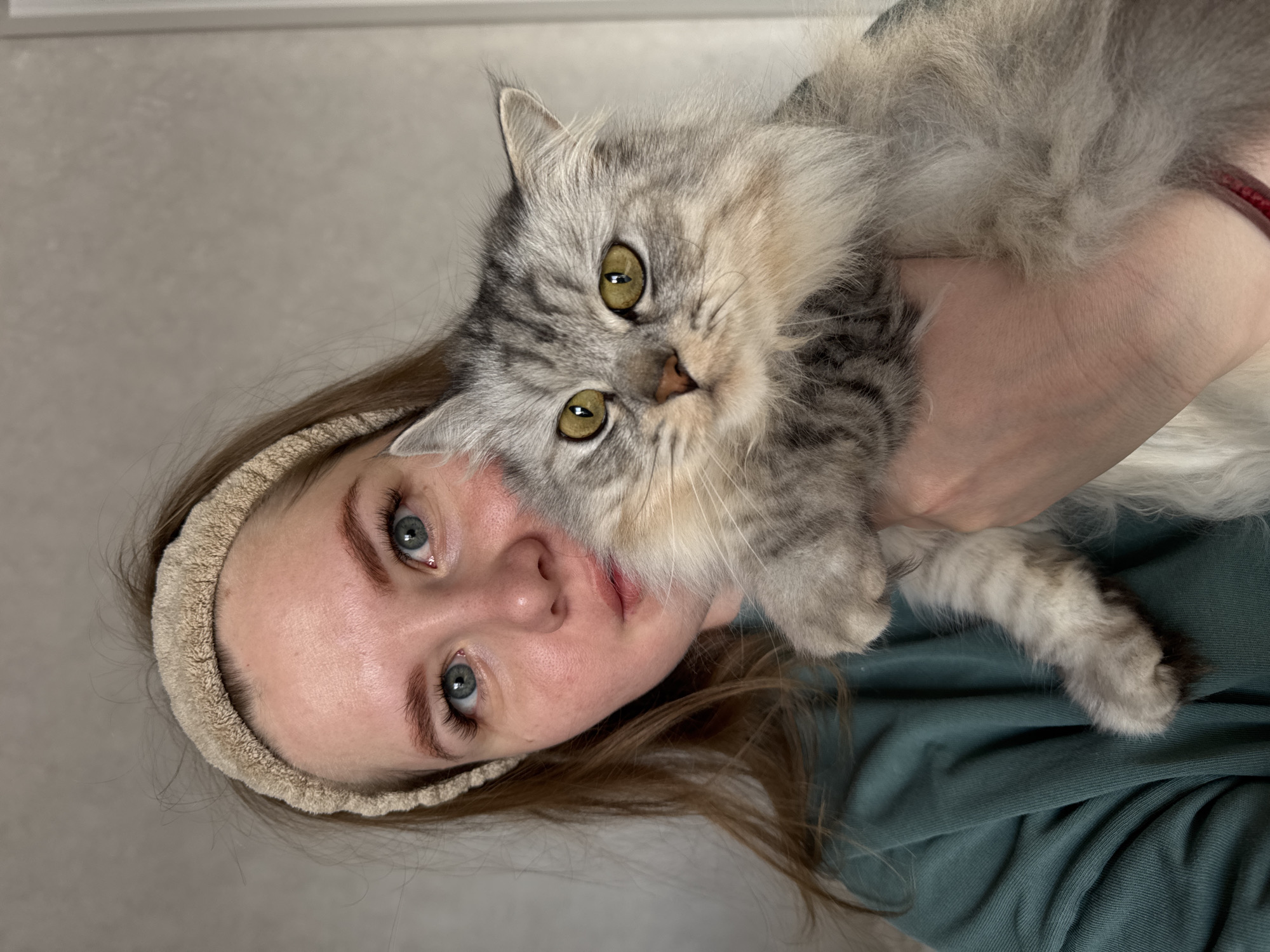 A woman with blue eyes and light brown hair, wearing a beige headband and a dark green shirt, is holding a fluffy, gray and white cat. The cat has yellow-green eyes and a gray striped pattern on its fur. The background appears to be a plain wall. The image orientation is rotated ninety degrees counter-clockwise. There is no indication of Gornyi (Dvurechenskoe S/P), Russia.