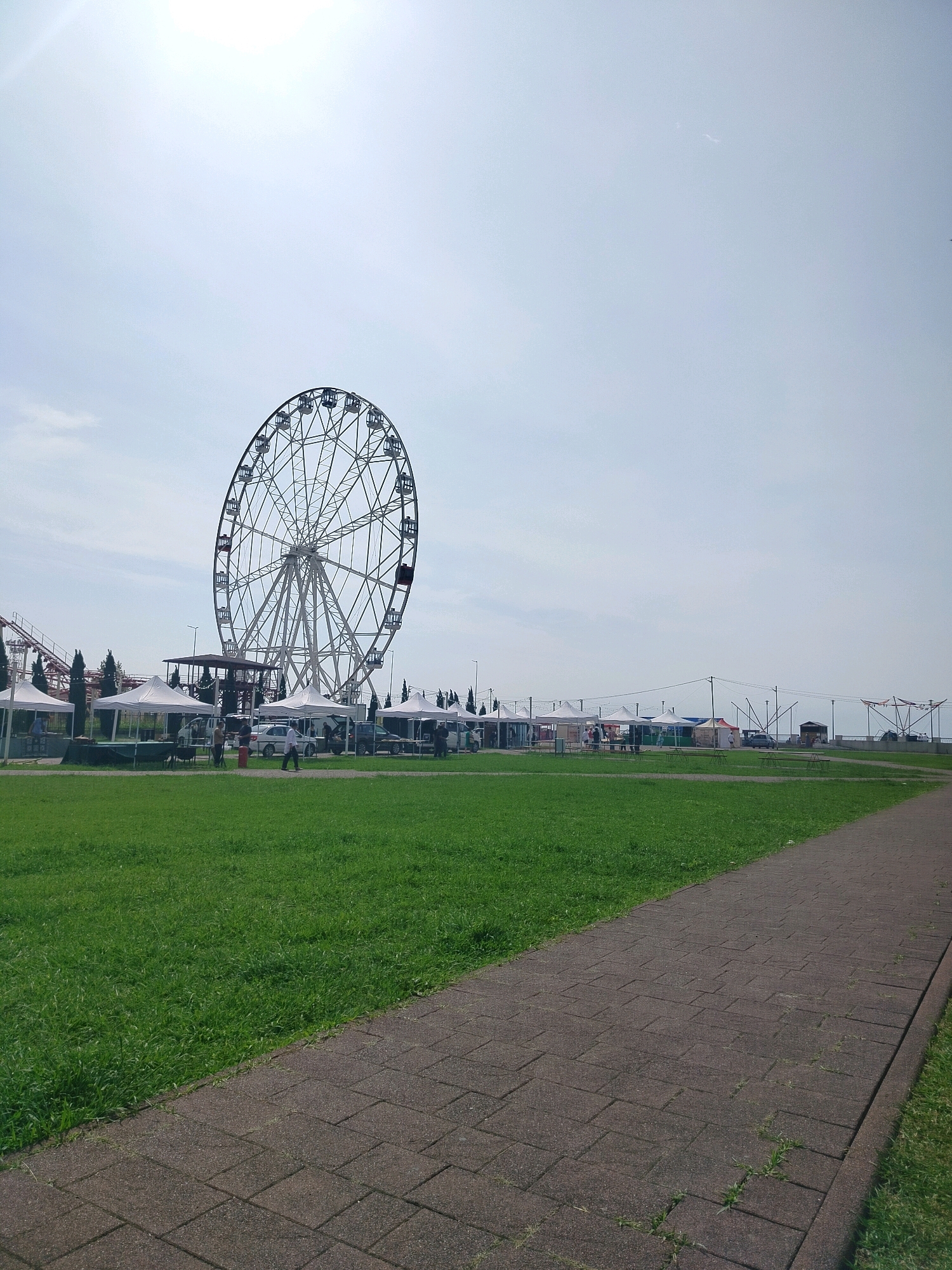 The image depicts an outdoor scene in Gagra, Abkhazia, featuring a large Ferris wheel as the prominent central element. The Ferris wheel is white with black cabins and is positioned on the left side of the frame. In front of the Ferris wheel, several white tents are set up, appearing to be market stalls or vendor booths. Several vehicles, including a white car and a dark-colored SUV, are parked near the tents. A roller coaster track is visible in the background on the far left. To the right of the Ferris wheel, a paved walkway runs diagonally from the lower right corner towards the center of the image, bordered by a strip of green grass. The sky is bright and overcast, with a large lens flare emanating from the upper left. The overall setting appears to be a recreational area or an amusement park.