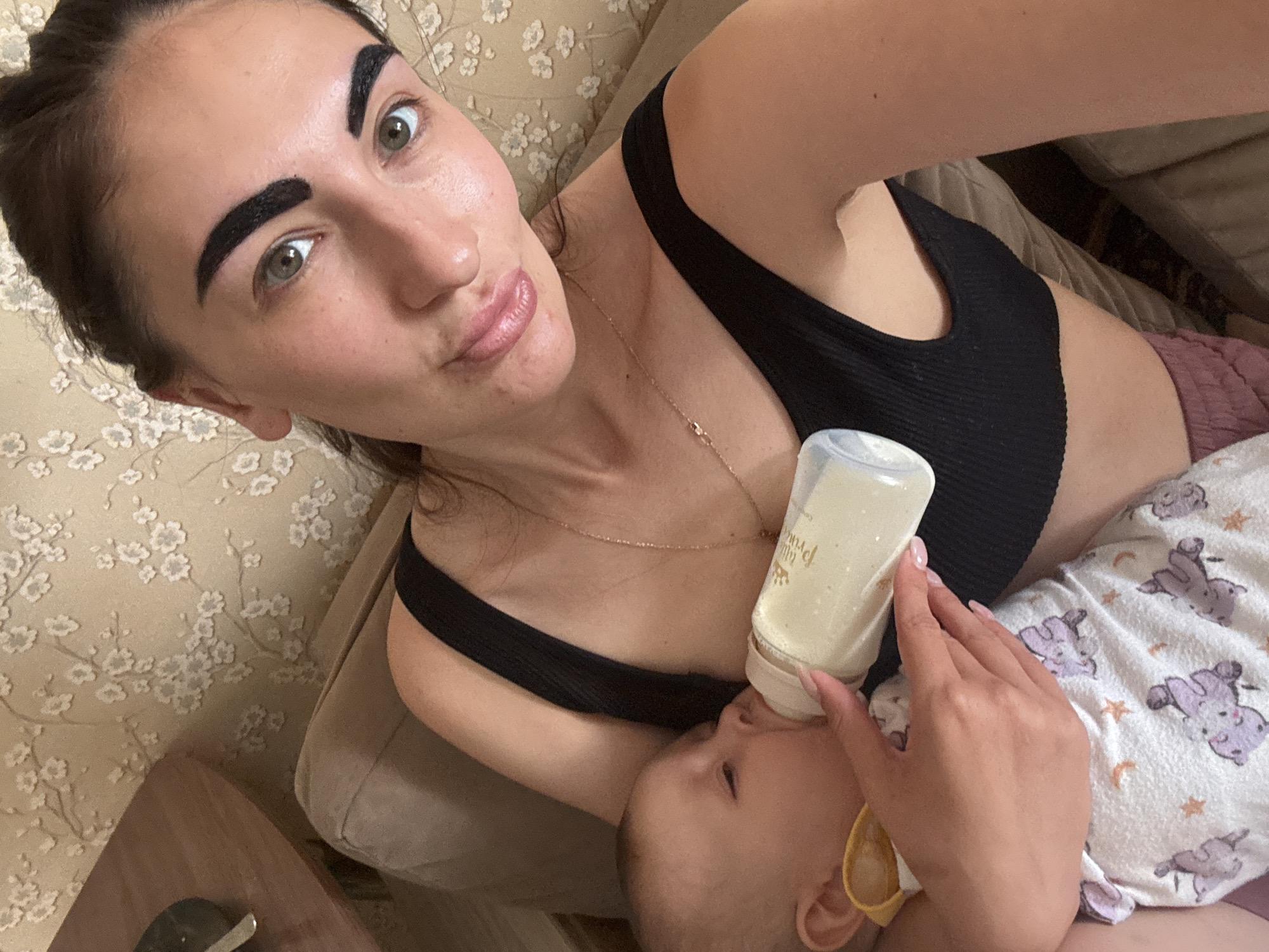 A woman is lying on a couch with a baby in her arms, feeding the baby from a bottle. The woman is fair-skinned with dark hair pulled back and thick, filled-in eyebrows. She is wearing a black ribbed tank top and a gold necklace. The baby is wearing a white onesie with a purple animal print. The bottle is filled with a white liquid. The couch has a beige floral pattern. There is a wooden table visible in the lower left corner of the image. The location is unknown; there is no discernible evidence indicating the city of Sochi, Russia.