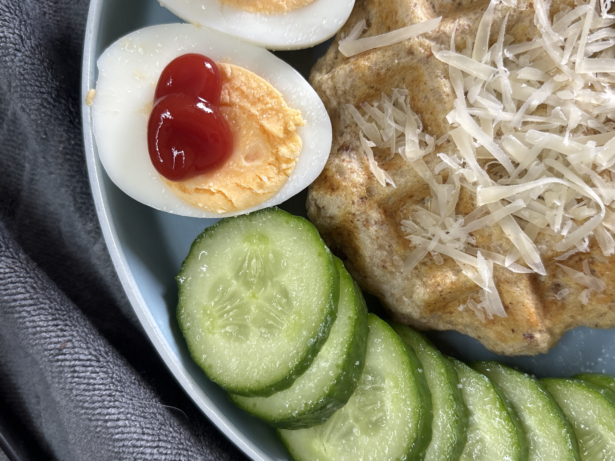 A close-up, overhead shot shows a light blue plate filled with a meal. On the left, there are several overlapping slices of cucumber. To the right of the cucumbers is a portion of what appears to be a waffle or pancake topped with shredded cheese. Adjacent to this is a hard-boiled egg, cut in half, with ketchup drizzled on the yolk. In the background, a gray, textured cloth is partially visible. There are no people or animals in the image, and no identifiable landmarks or specific locations are present.
