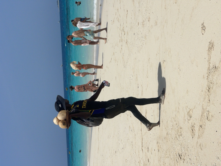 A sunny beach scene unfolds in Zanzibar, Tanzania, featuring light-colored sand, clear turquoise ocean water, and a bright blue sky. In the foreground, a dark-skinned male vendor walks barefoot from left to right, wearing a black wide-brimmed hat, a dark blue and yellow t-shirt with "TANZAN" visible, black athletic pants with "NIKE" on the left leg, and a grey backpack. He balances a circular wooden tray holding three coconuts on his right shoulder and holds a small white object in his right hand. His feet are covered in sand, and a distinct shadow is cast to his right.
In the midground, several individuals are visible on the sand and in the shallow water. To the right of the vendor, a blonde woman in a red bikini stands, looking at a mobile device. Behind her, two other women in bikinis stand. Further right, closer to the water's edge, a male in light shorts walks left, another male in darker shorts stands, and a woman in a white cover-up wades in the water. Distant figures are visible in the ocean. The water exhibits small waves at the shore, and several small boats, including what appears to be a dhow, are visible further out on the water under a clear sky.