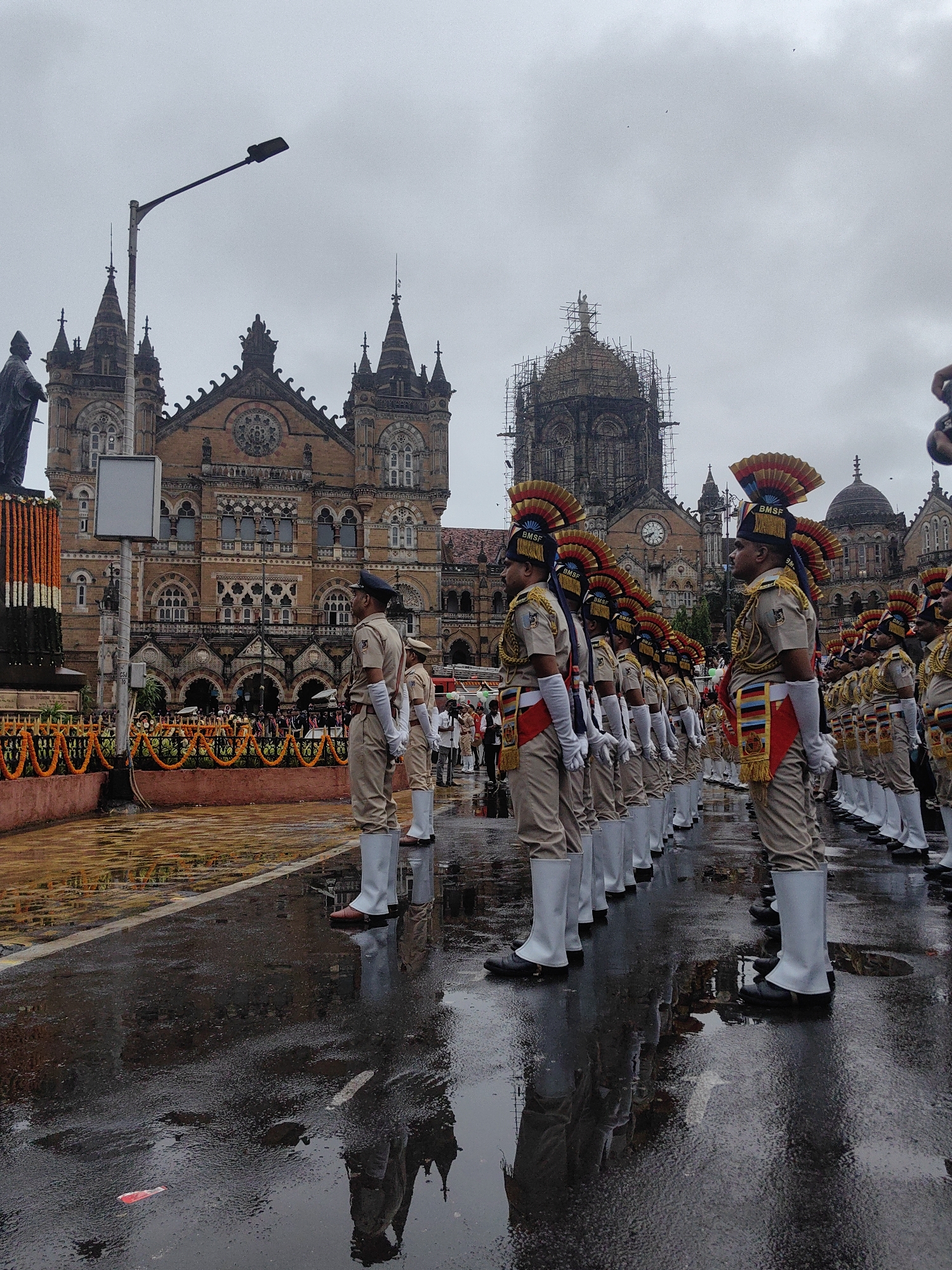 A line of men in formal uniforms, some with decorative headwear and sashes, stand at attention on a wet, reflective street. They appear to be participating in a parade or ceremony. Behind them, a grand, ornate building with intricate architecture and multiple spires looms under a cloudy sky. The building, characterized by its arched windows and decorative stonework, is a prominent landmark. One of the spires is partially covered in scaffolding. The street itself is covered in puddles, reflecting the surrounding structures and the overcast sky. In the foreground, a statue stands on a platform decorated with orange and white garlands. This scene is indicative of a ceremonial event taking place in front of the Chhatrapati Shivaji Maharaj Terminus, a historic railway station and UNESCO World Heritage Site in Mumbai, India.